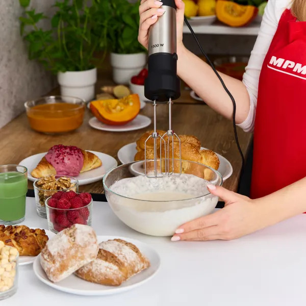 Osoba używa miksera elektrycznego do ubijania śmietany. Różne wypieki, owoce i warzywa ułożone na stole.
