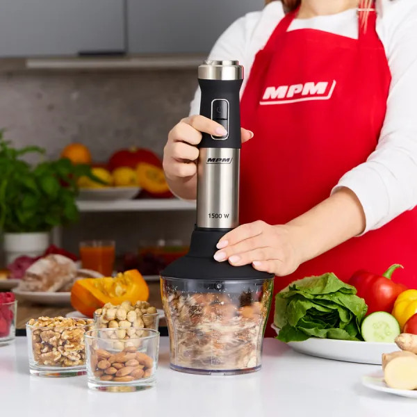 Osoba używająca srebrno-czarnego blendera do siekania jedzenia. Na stole znajdują się inne składniki, takie jak orzechy, warzywa i owoce.