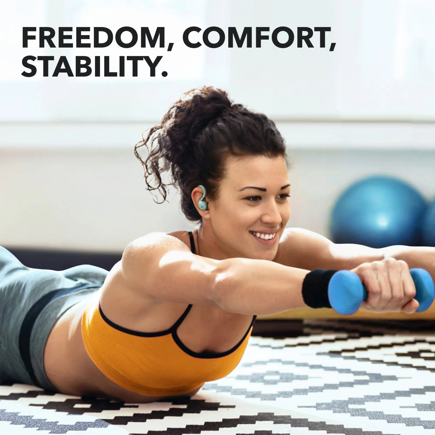 Kobieta ćwiczy z hantlami, w żółtym topie i szarych spodniach, z turkusowymi słuchawkami. W tle obecne są niebieskie piłki.