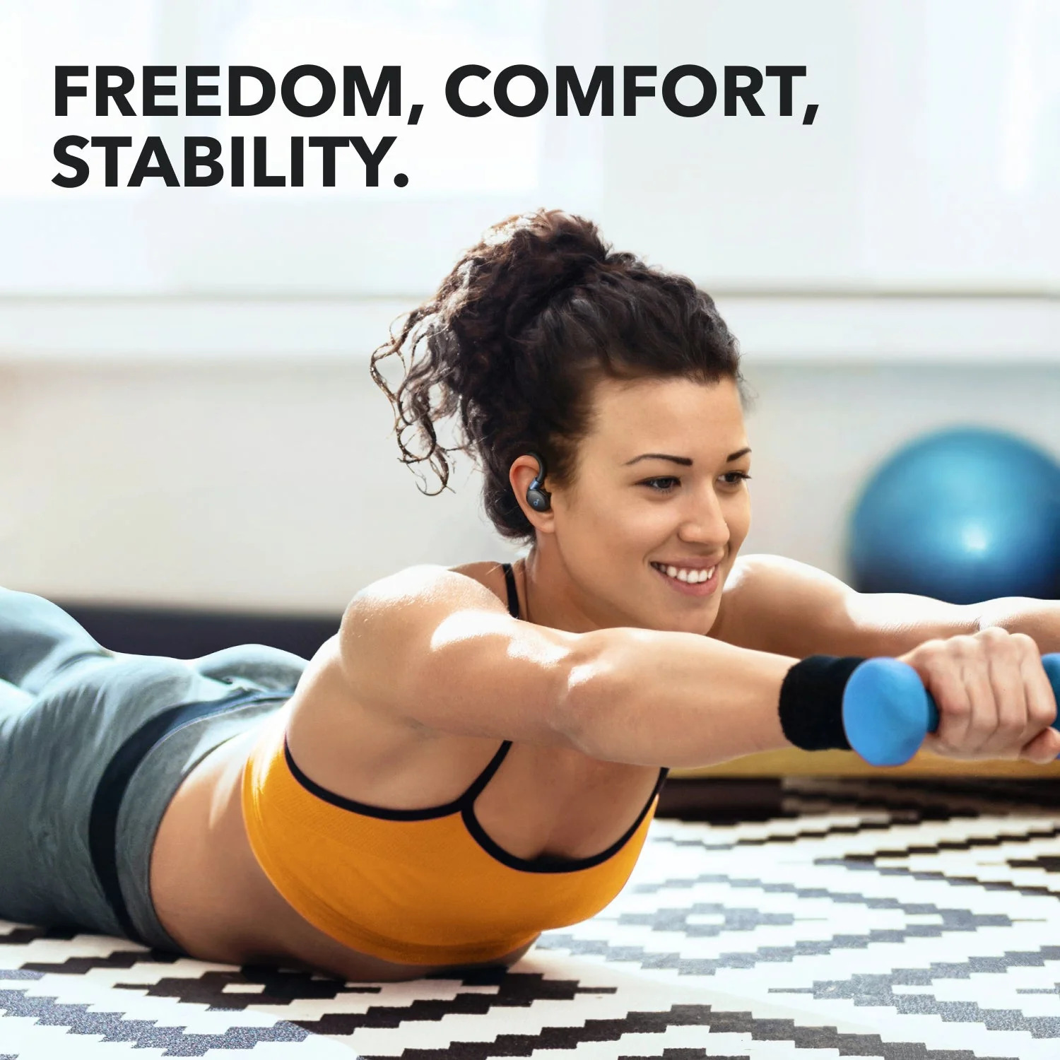 Kobieta w żółtym topie ćwiczy z niebieskimi ciężarkami, nosząc słuchawki. Czarno-biały wzorzysty dywan, niebieska piłka gimnastyczna w tle.