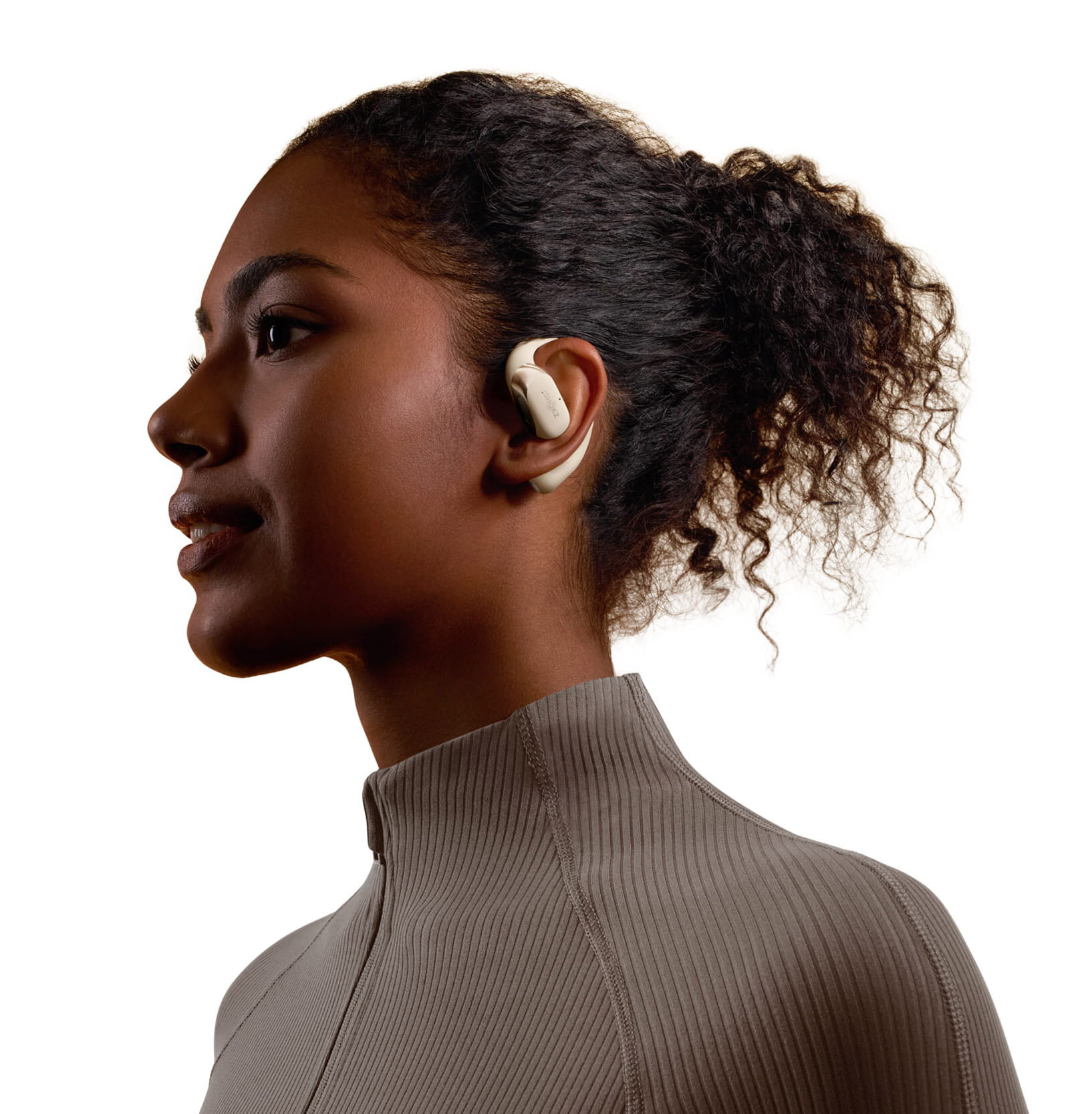 Kobieta o ciemnej skórze nosząca beżową słuchawkę. Ma ciemne kręcone włosy i szarą górę.