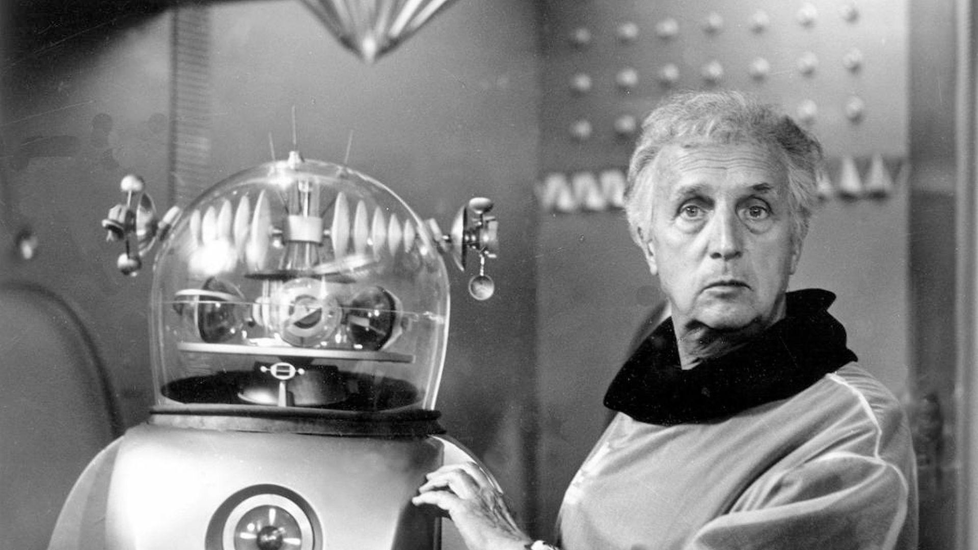 Ein Mann in einem hellen Hemd steht neben einem Retro-Roboter. Der Roboter hat einen durchsichtigen Kuppelkopf. Beide schauen in die Kamera.