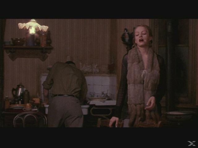 Eine Frau mit Pelzstola steht in der Nähe eines Mannes an einem Waschbecken. Eine Lampe und Regale sind sichtbar. Küchenszene.