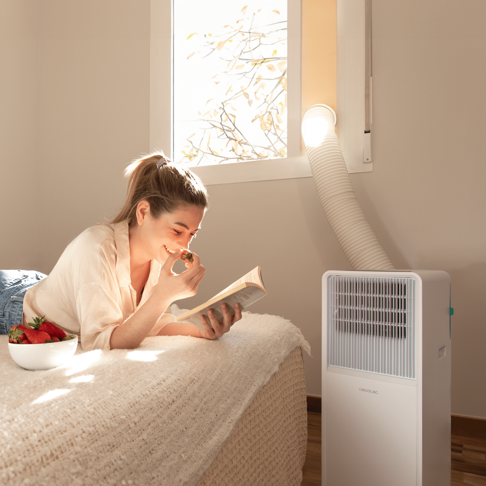 Eine Frau liegt auf einem Bett, liest und isst eine Erdbeere. Klimaanlage daneben. Fenster im Hintergrund.