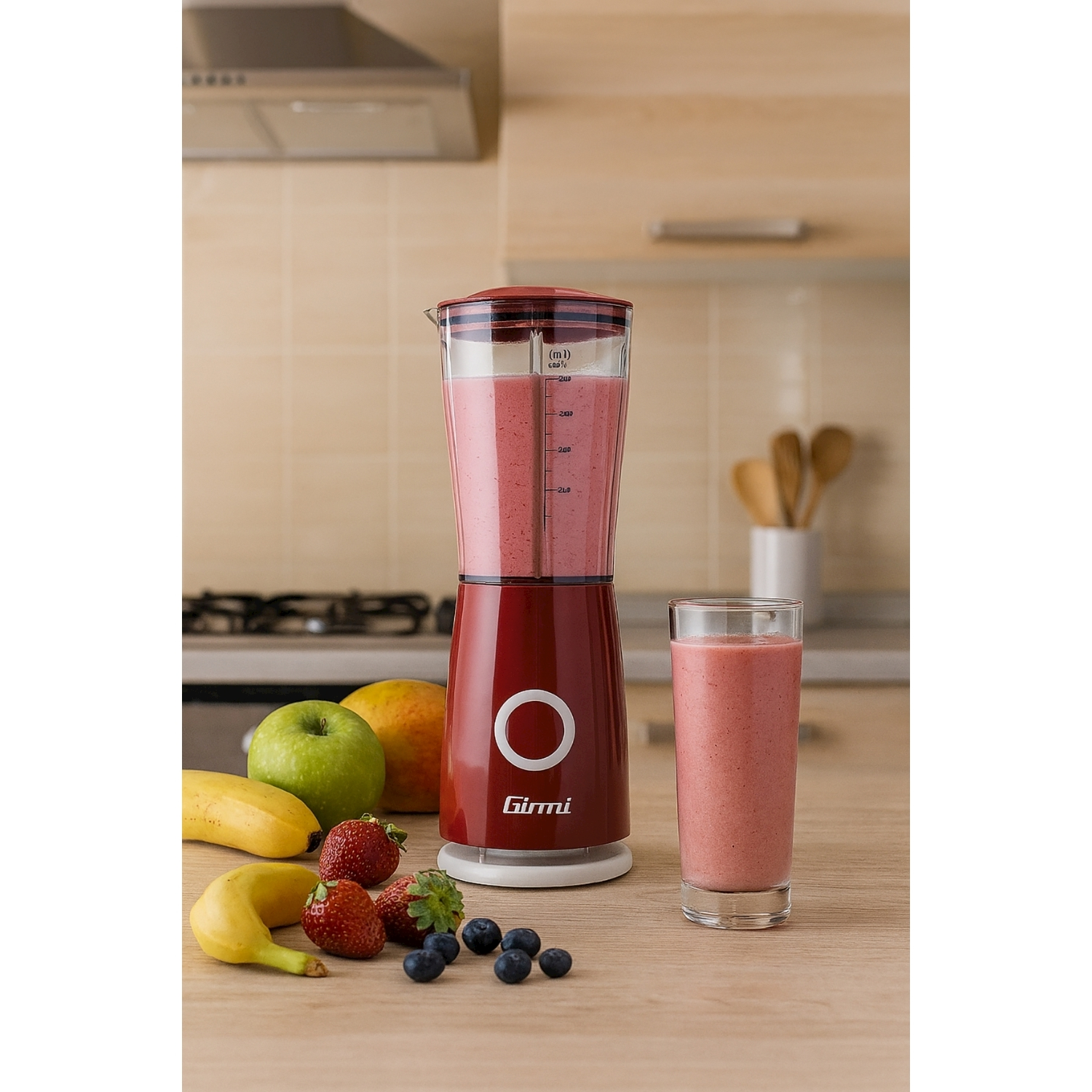 Un frullatore rosso con frullato, frutta (banane, fragole, mela, mango e mirtilli) e un bicchiere di frullato su un tavolo di legno.