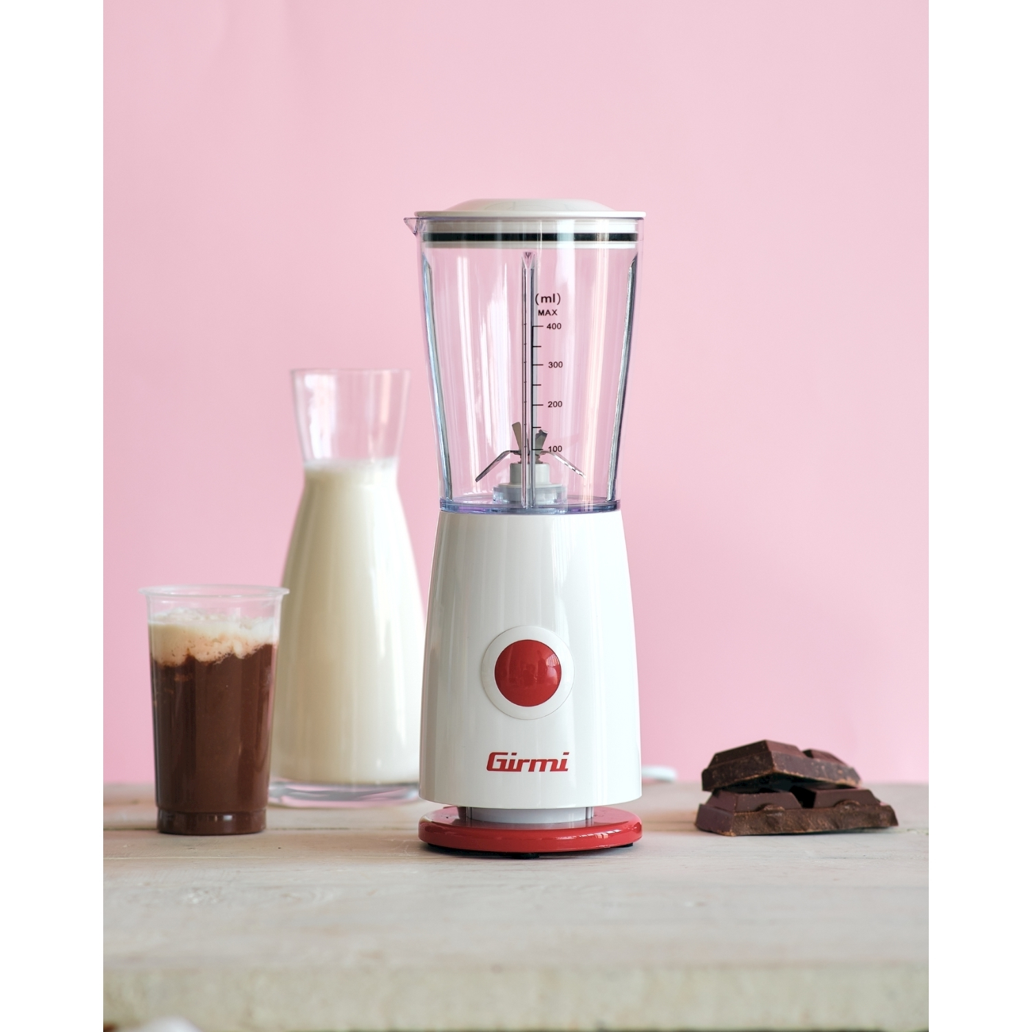 Un frullatore bianco con pulsante e base rossi, accanto a un bicchiere di cioccolata, una caraffa di latte e pezzi di cioccolato.