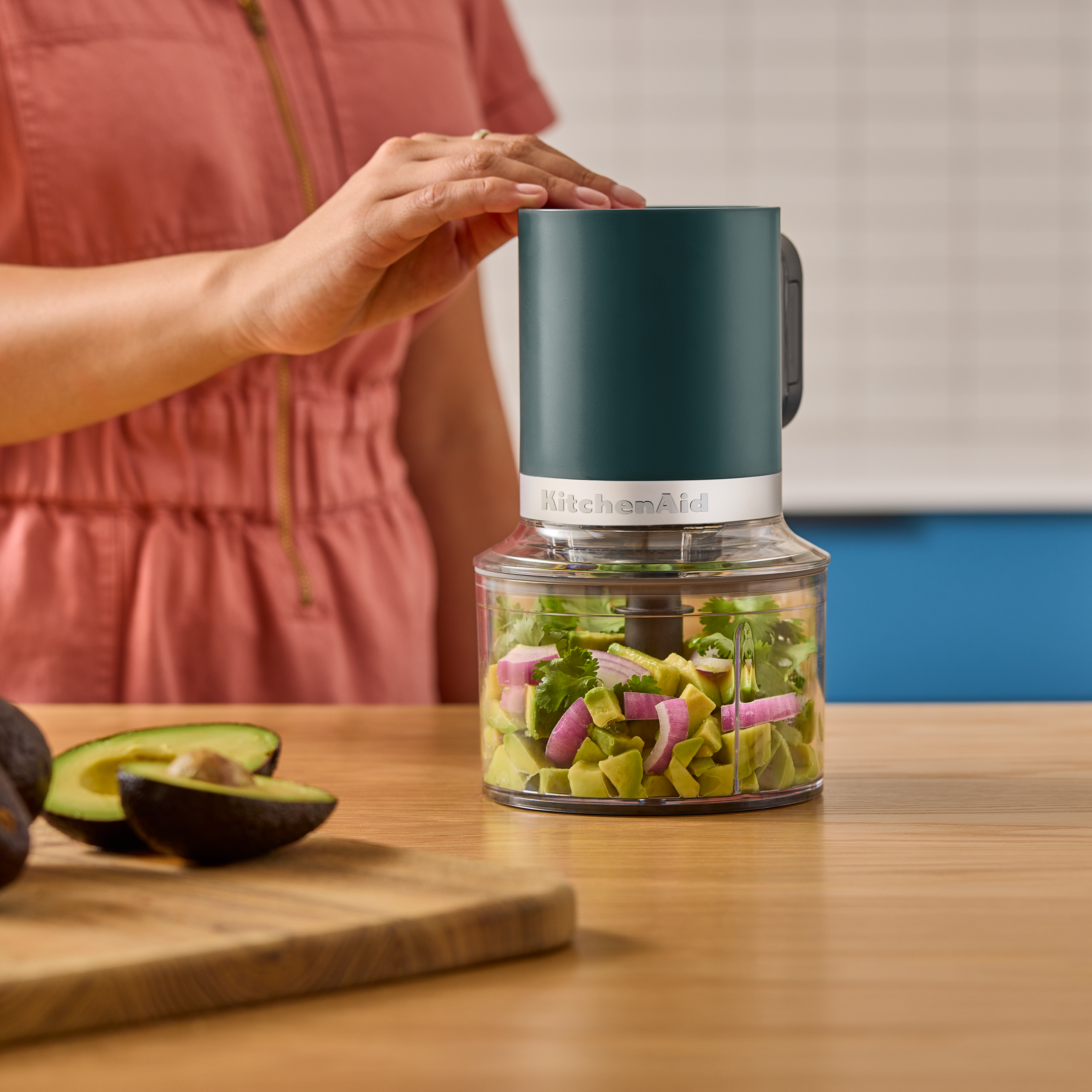 Die Hand einer Person liegt auf einem grünen KitchenAid-Zerkleinerer. Er ist mit gewürfelten Avocados, roten Zwiebeln und Koriander gefüllt.