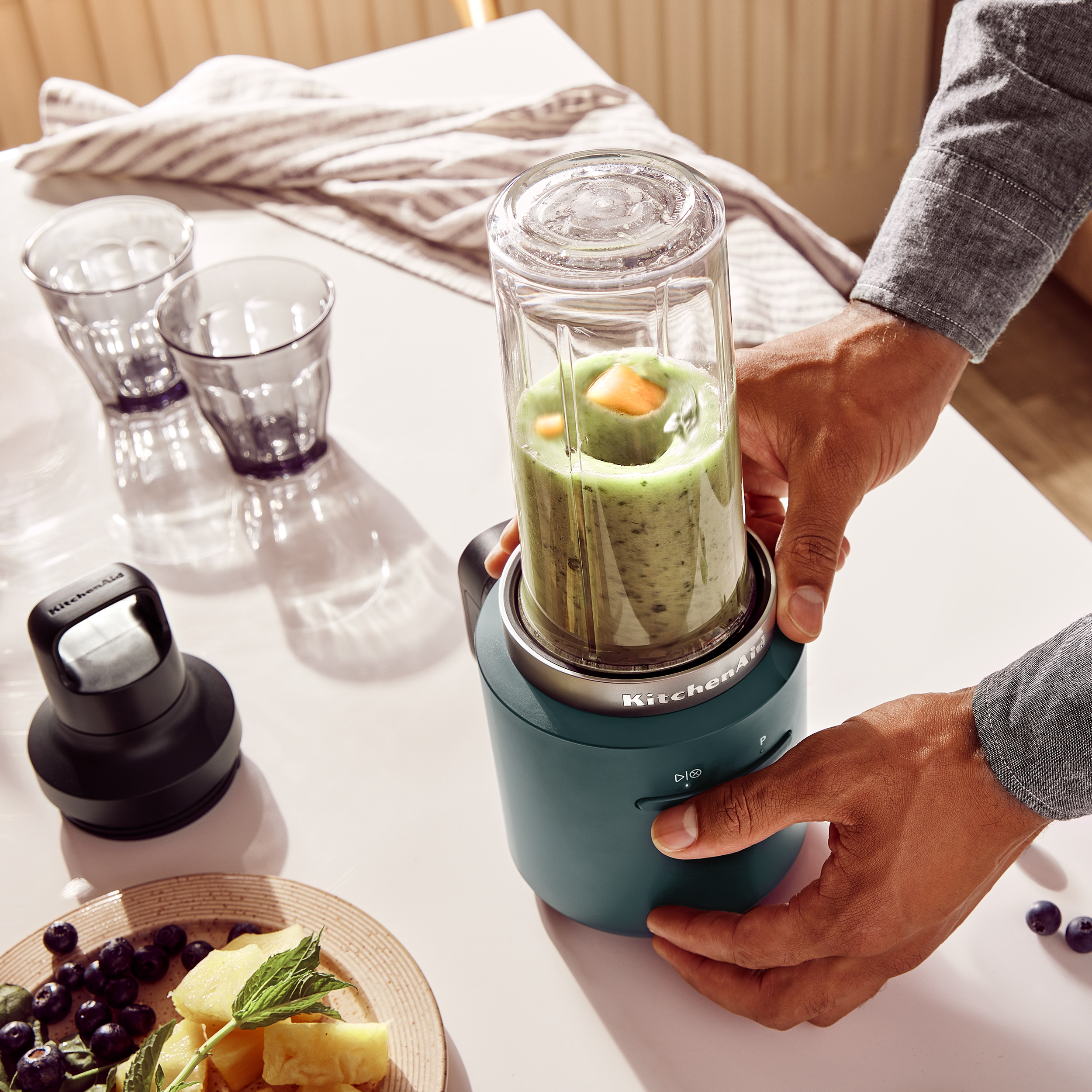 Eine Person bereitet einen Smoothie mit einem Mixer zu. Auf dem Tisch liegen Früchte und Gläser. Der Mixer ist teal.