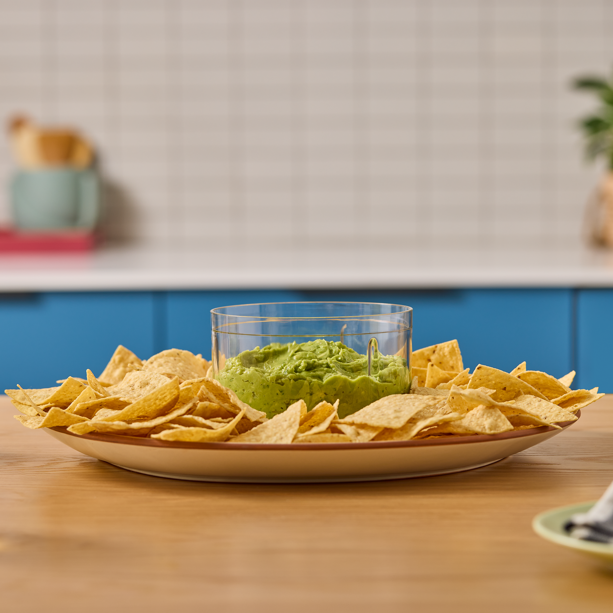 Eine Schüssel Guacamole ist von Tortilla-Chips auf einem Holztisch umgeben. Hintergrund zeigt Küchenelemente.