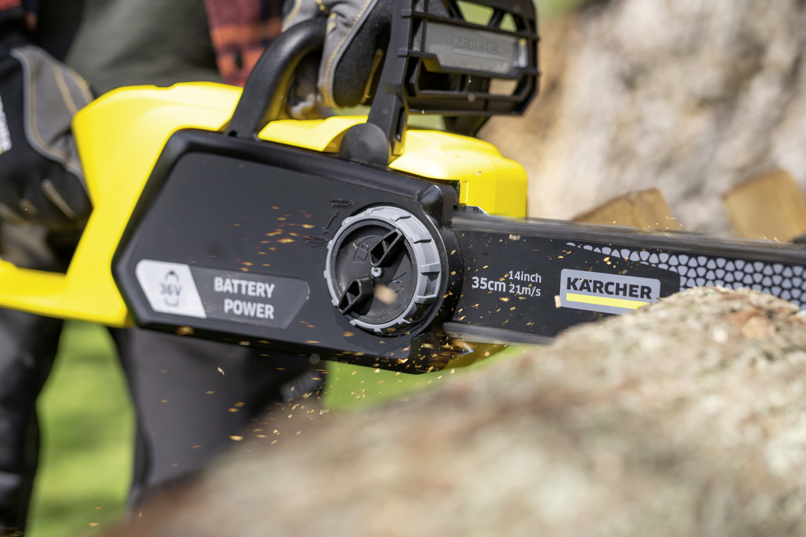 Egy sárga-fekete Karcher láncfűrész fát vág. Fűrészpor repül. A fűrészen a 'Battery Power' felirat látható.