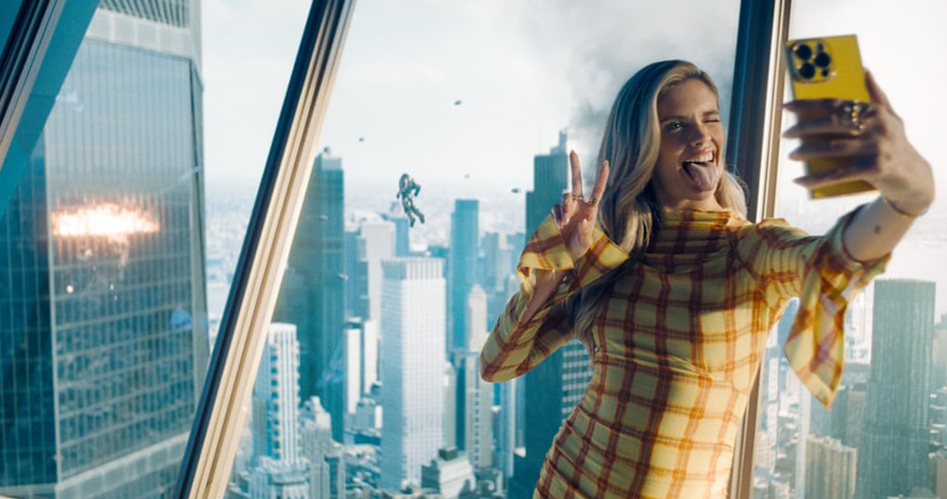 Frau macht Selfie in gelbem Kleid mit Stadtkulisse. Eine Figur fliegt vor dem Fenster.