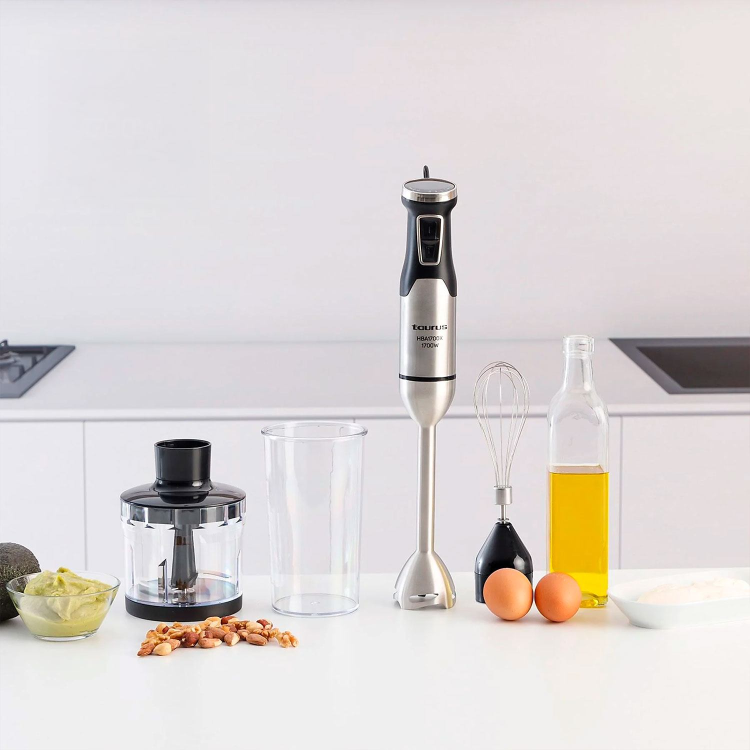 Escena de cocina con batidora, ingredientes y herramientas. Incluye aguacate, nueces, huevos, aceite y un batidor sobre superficie blanca.