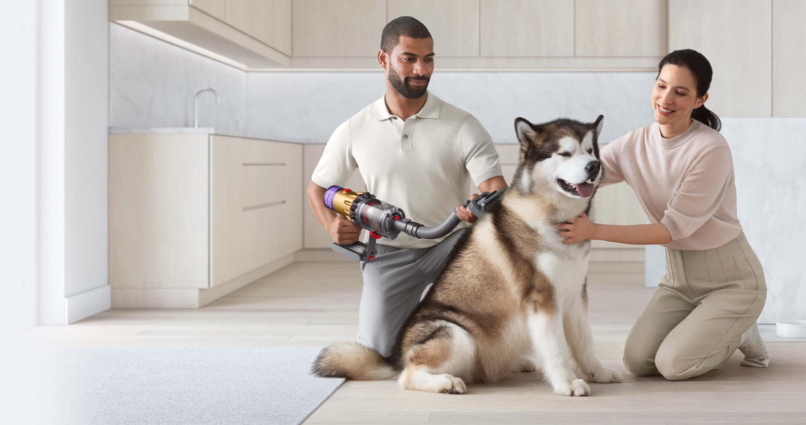Un hombre aspira a un perro grande con una Dyson. Una mujer se arrodilla a su lado. Cocina moderna al fondo.