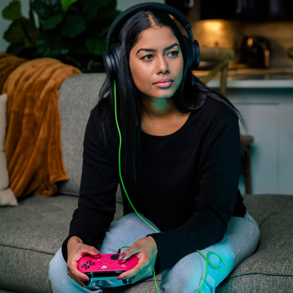 Frau mit Headset und rosa Gamecontroller, sitzt auf einem Sofa. Grünes Kabel verbindet Controller und Headset.