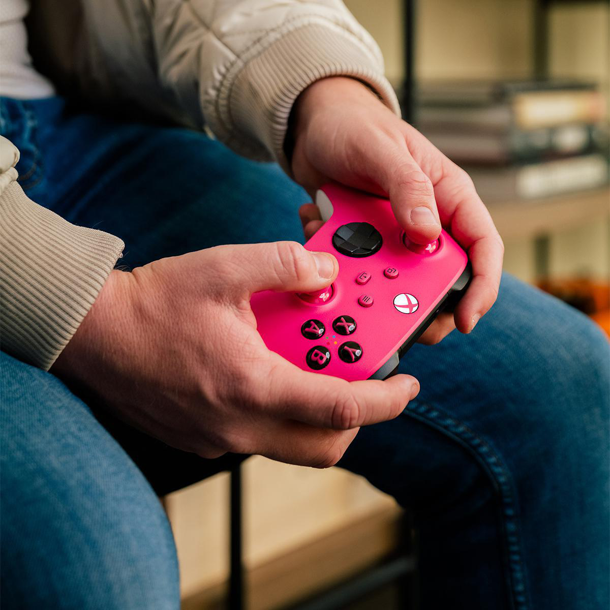 Eine Person hält einen pinkfarbenen Videospiel-Controller. Die Person trägt eine cremefarbene Jacke und Jeans und sitzt. Die Controller-Tasten sind schwarz.