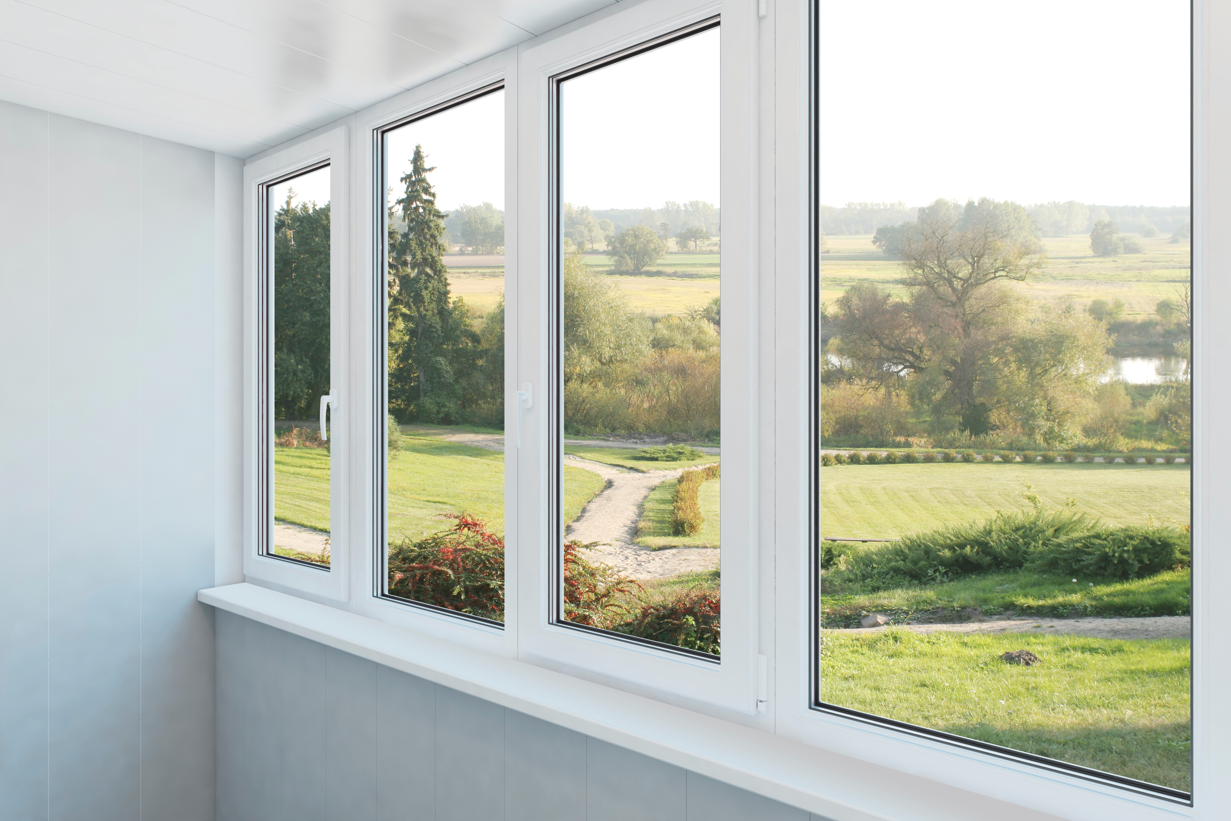 Blick durch ein Fenster. Draußen gibt es grünes Gras, Bäume, einen Weg und ein Gewässer. Drinnen gibt es eine weiße Fensterbank und Wand.