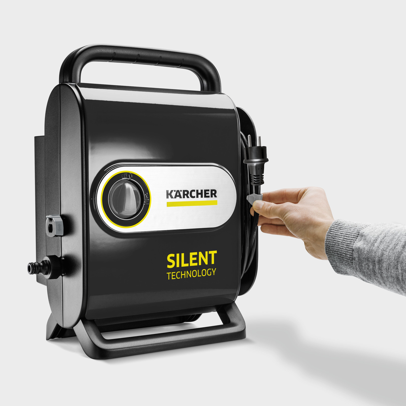 Fekete Karcher nagynyomású mosó, egy kéz bedugja a tápkábelt. Silent Technology. Fehér háttér.