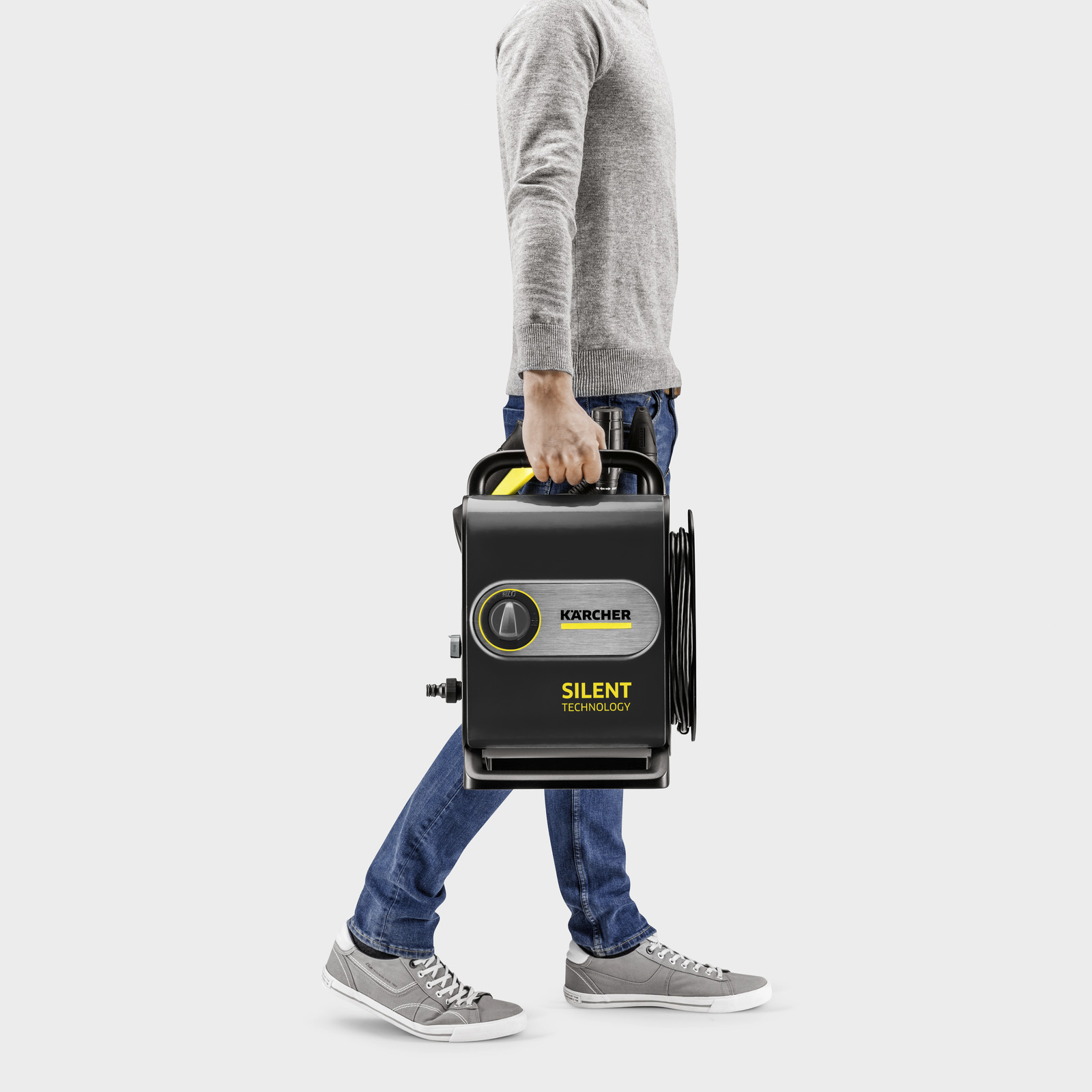 Egy személy egy fekete Kärcher nagynyomású mosót visz, jobbra sétál. A mosó sárga díszítéssel és a 'SILENT TECHNOLOGY' felirattal rendelkezik.