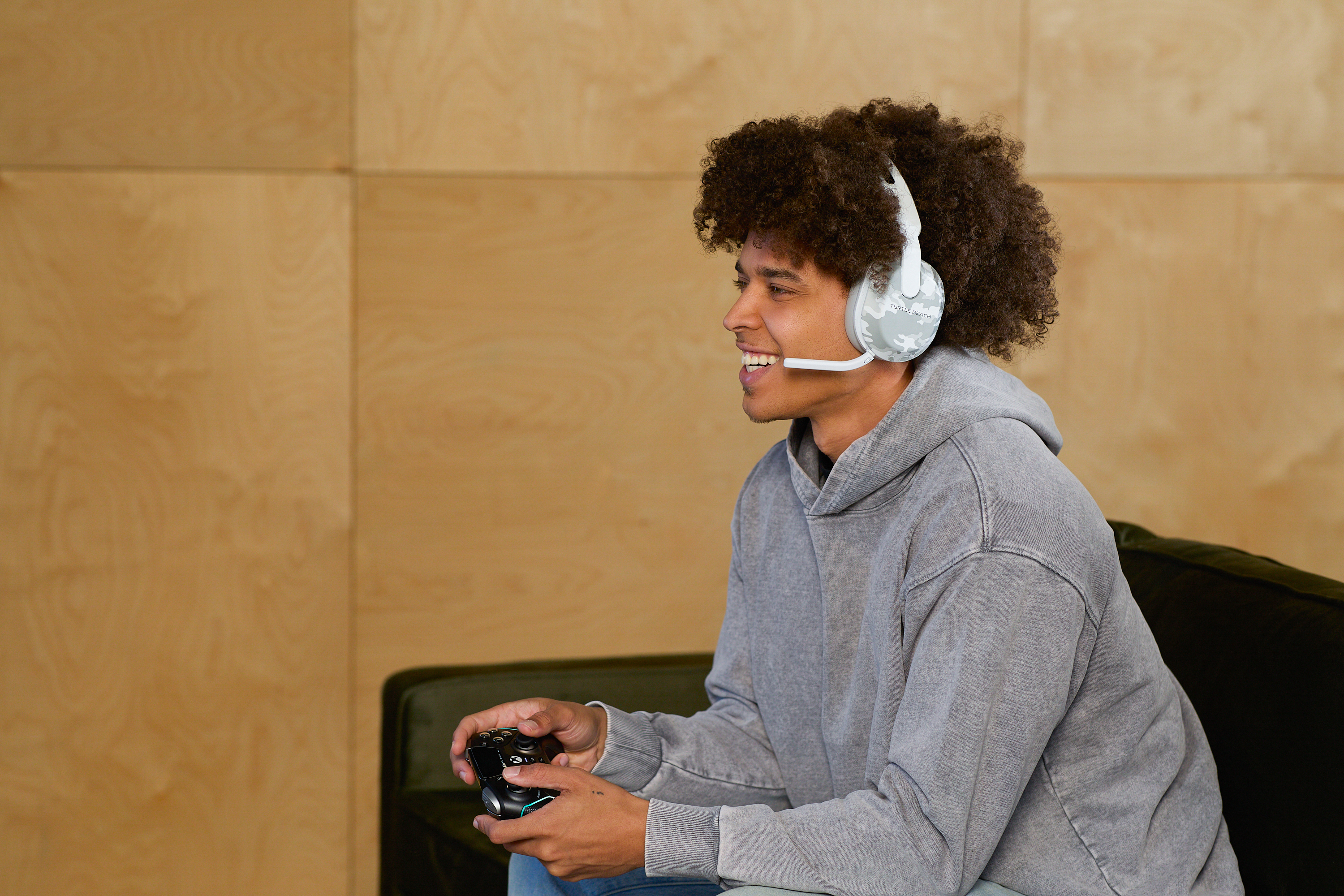 Ein Mann mit Kopfhörern und einem Controller lächelt und sitzt auf einem Sofa. Holzpaneele im Hintergrund.