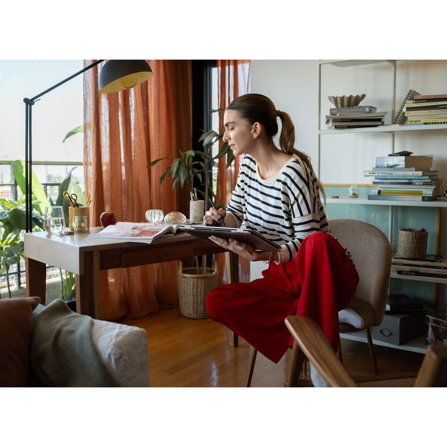Woman in striped top and red pants works on a tablet. The scene includes a wooden table, curtains, and a bookshelf in the background.