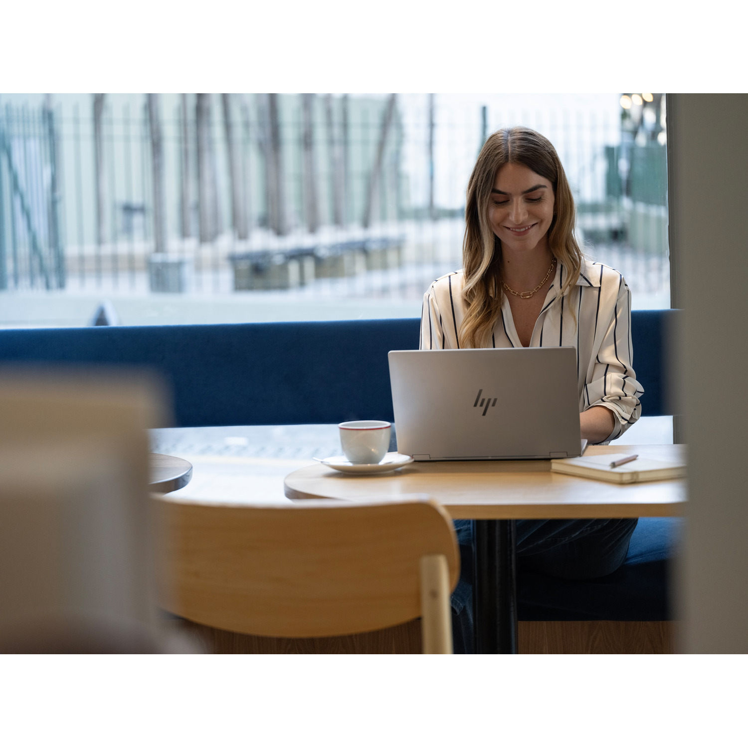 A woman smiles while using a laptop at a table. A coffee cup and notebook are on the table. Background shows windows and blue seating.
