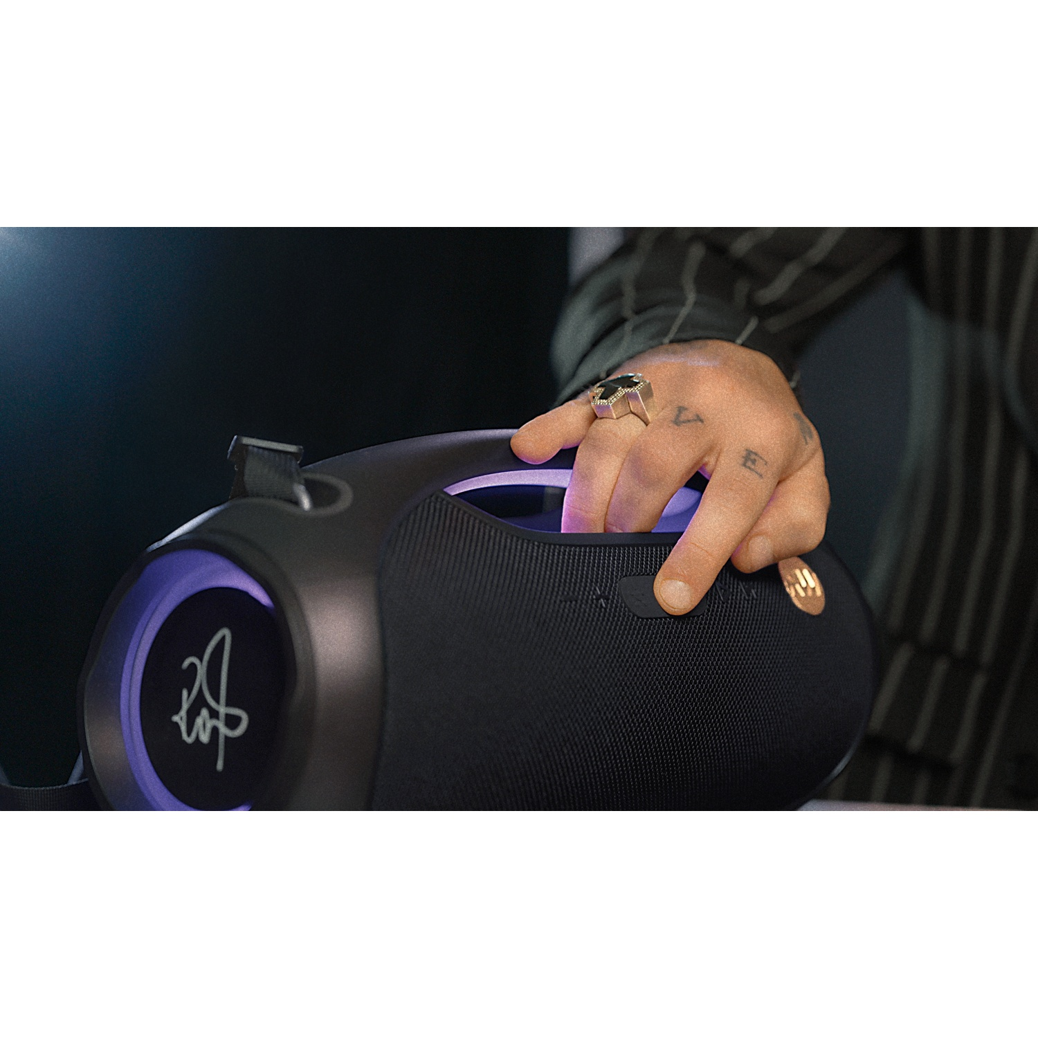 Una mano tiene in mano un altoparlante nero. La mano ha tatuaggi e un anello. L'altoparlante ha un logo e luci viola.