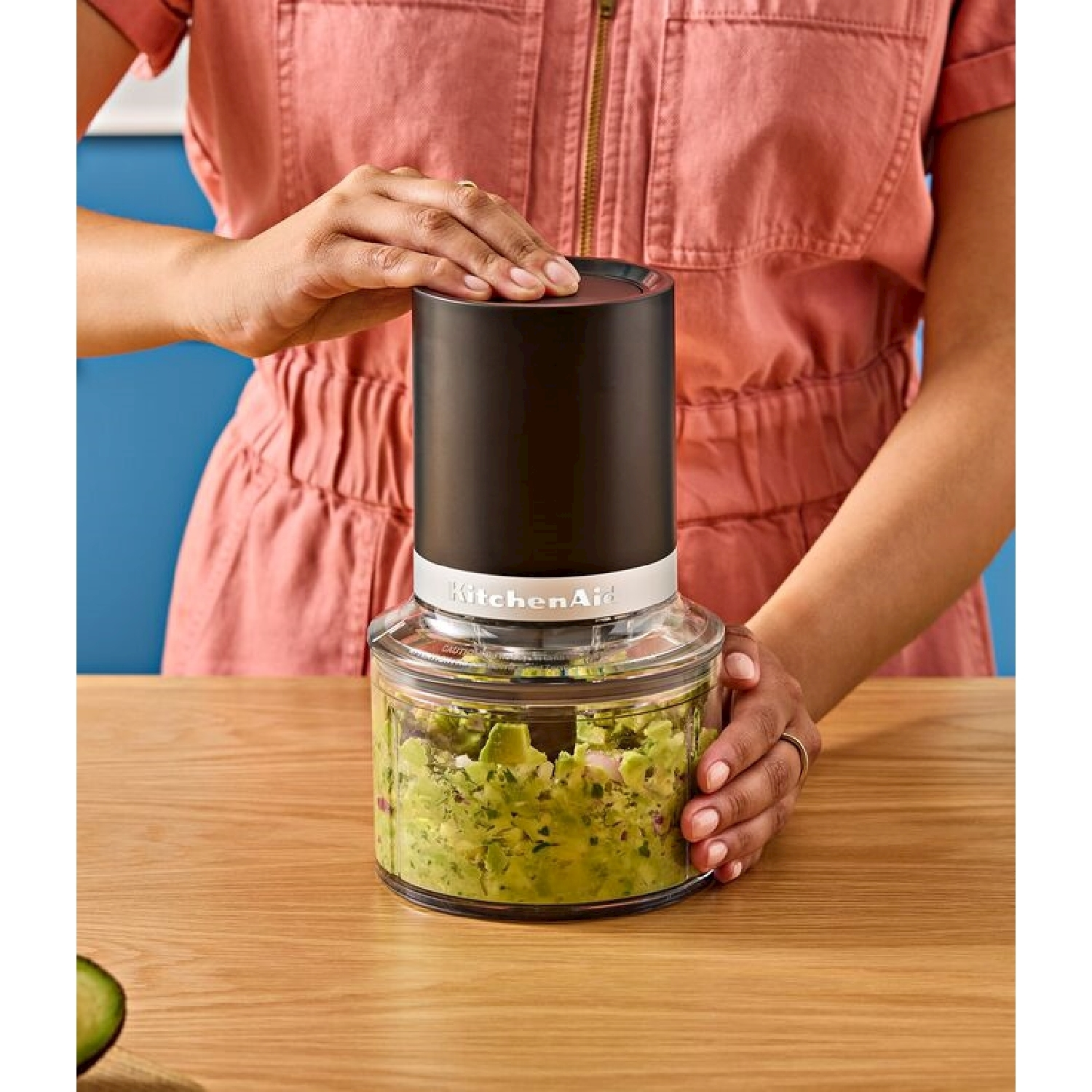 Una persona tiene in mano un robot da cucina nero e trasparente. Il robot contiene avocado tritato. La persona indossa una tuta rosa.