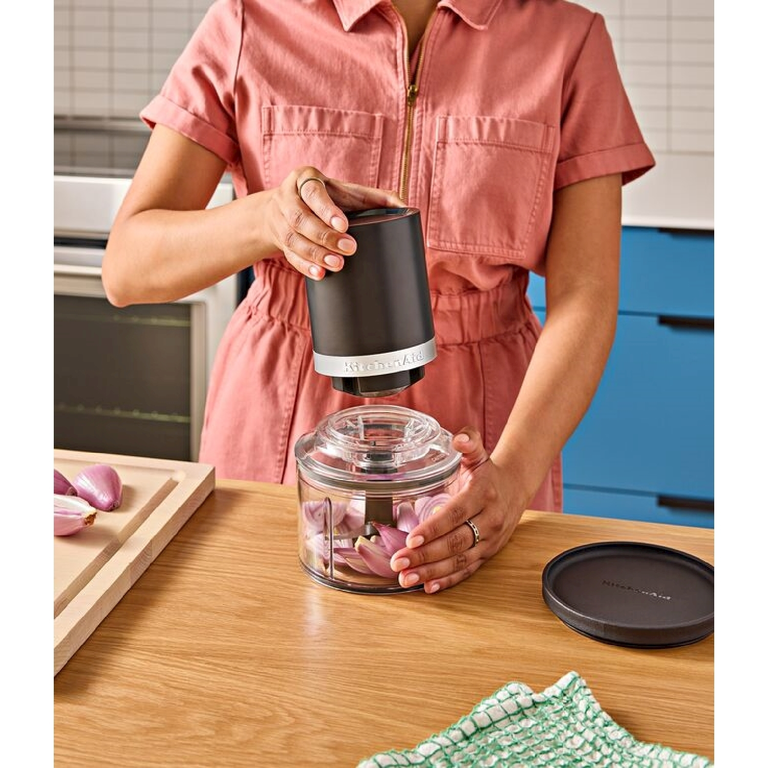 Una donna in tuta rosa usa un tritatutto KitchenAid. Scalogni sono nel tritatutto e su un tagliere su un tavolo di legno.
