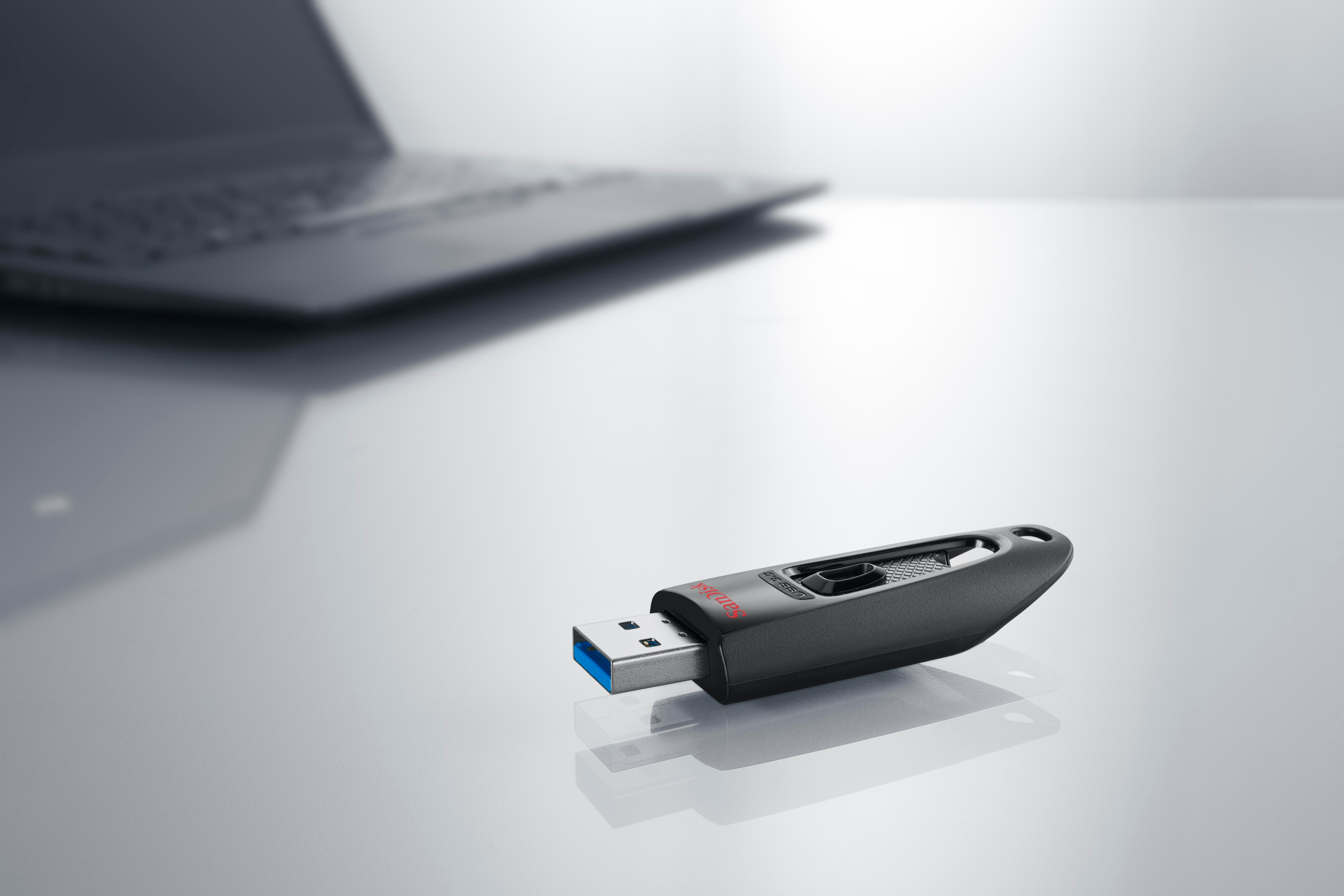 Ein schwarzer USB-Stick liegt auf einer reflektierenden Oberfläche. Ein Laptop ist unscharf im Hintergrund.