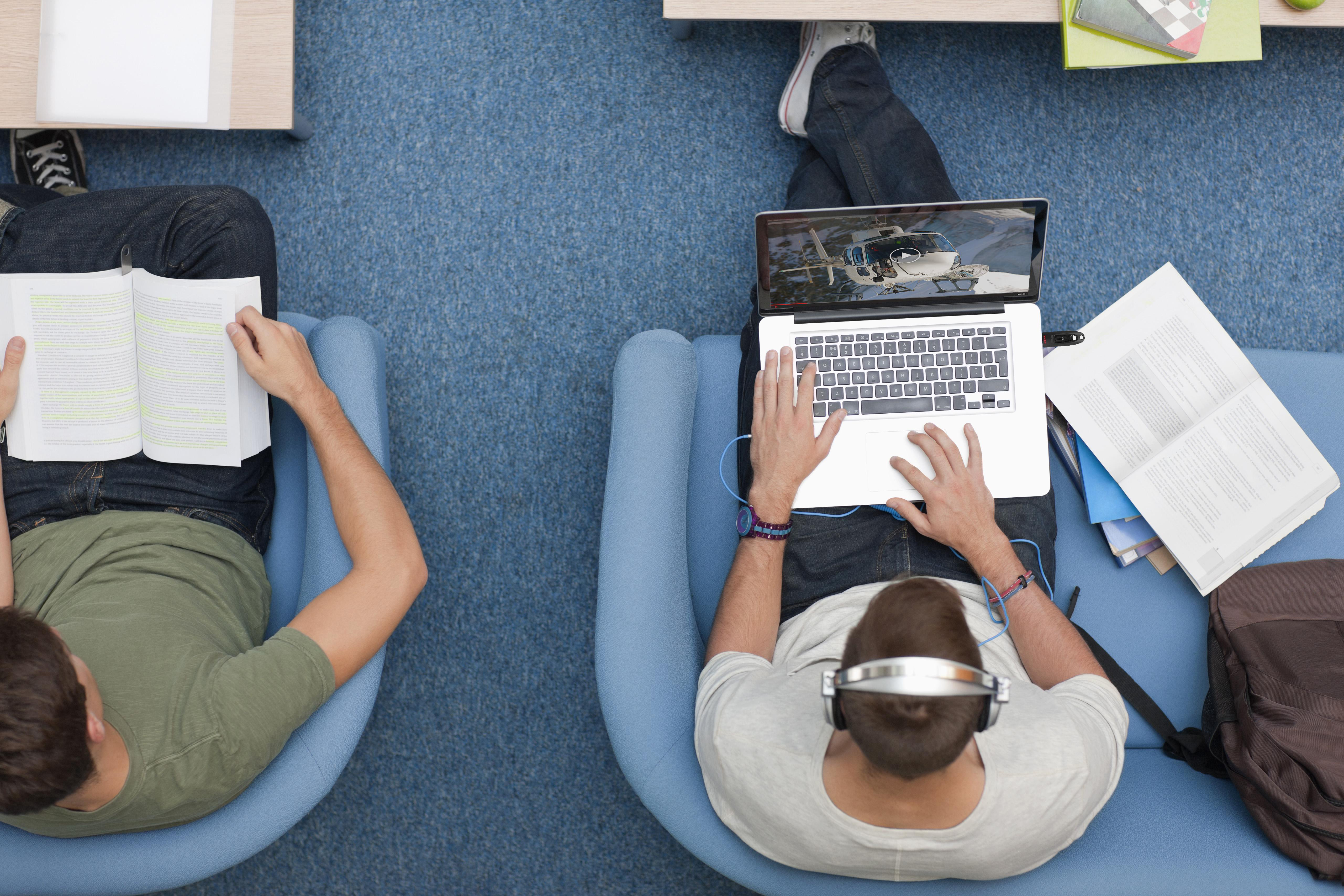 Zwei Personen in Stühlen, eine liest ein Buch, die andere benutzt einen Laptop mit Kopfhörern. Blauer Teppich und ein Tisch sind sichtbar.
