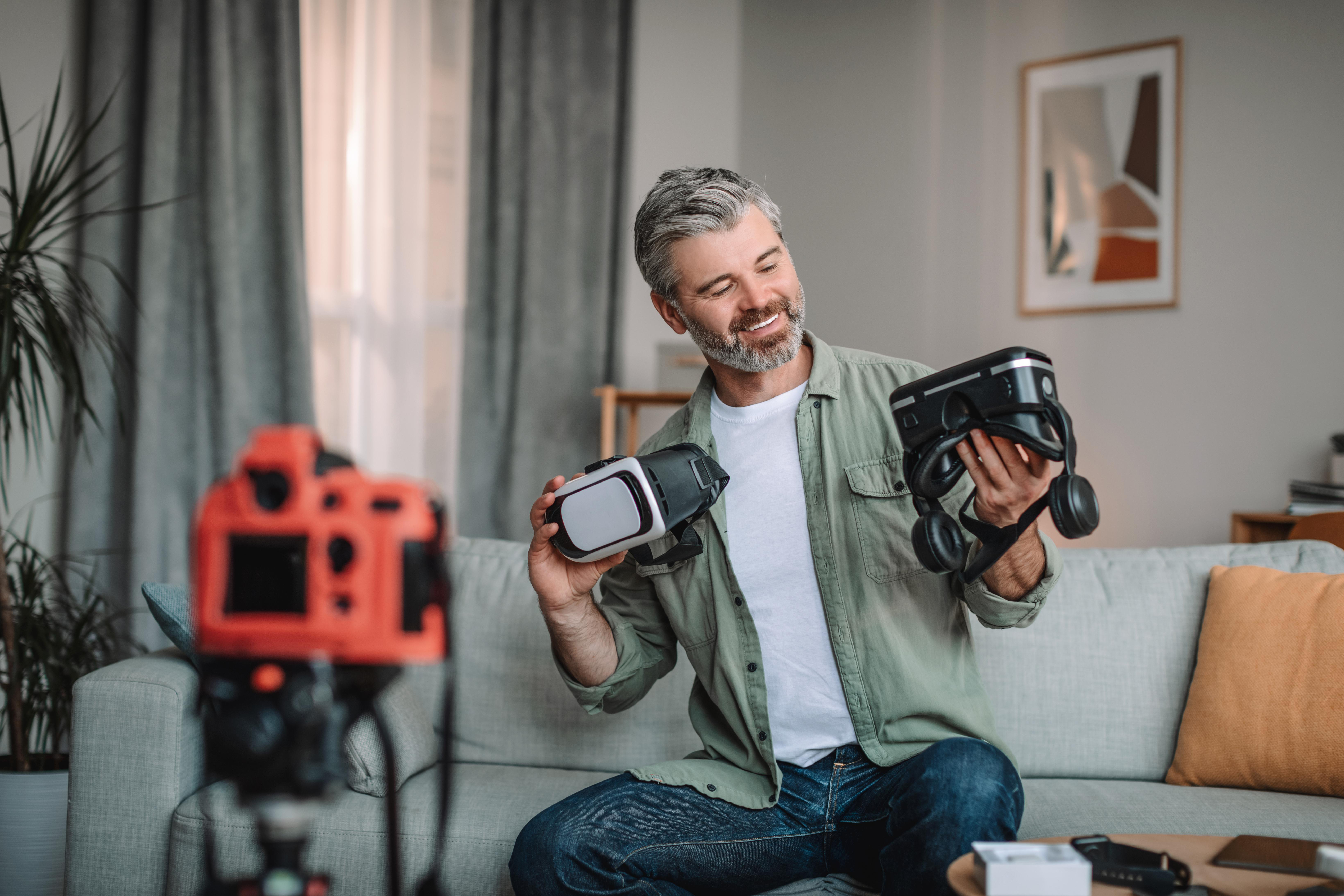 Ein Mann hält VR-Headsets. Er sitzt auf einem Sofa. Eine rote Kamera befindet sich im Vordergrund.