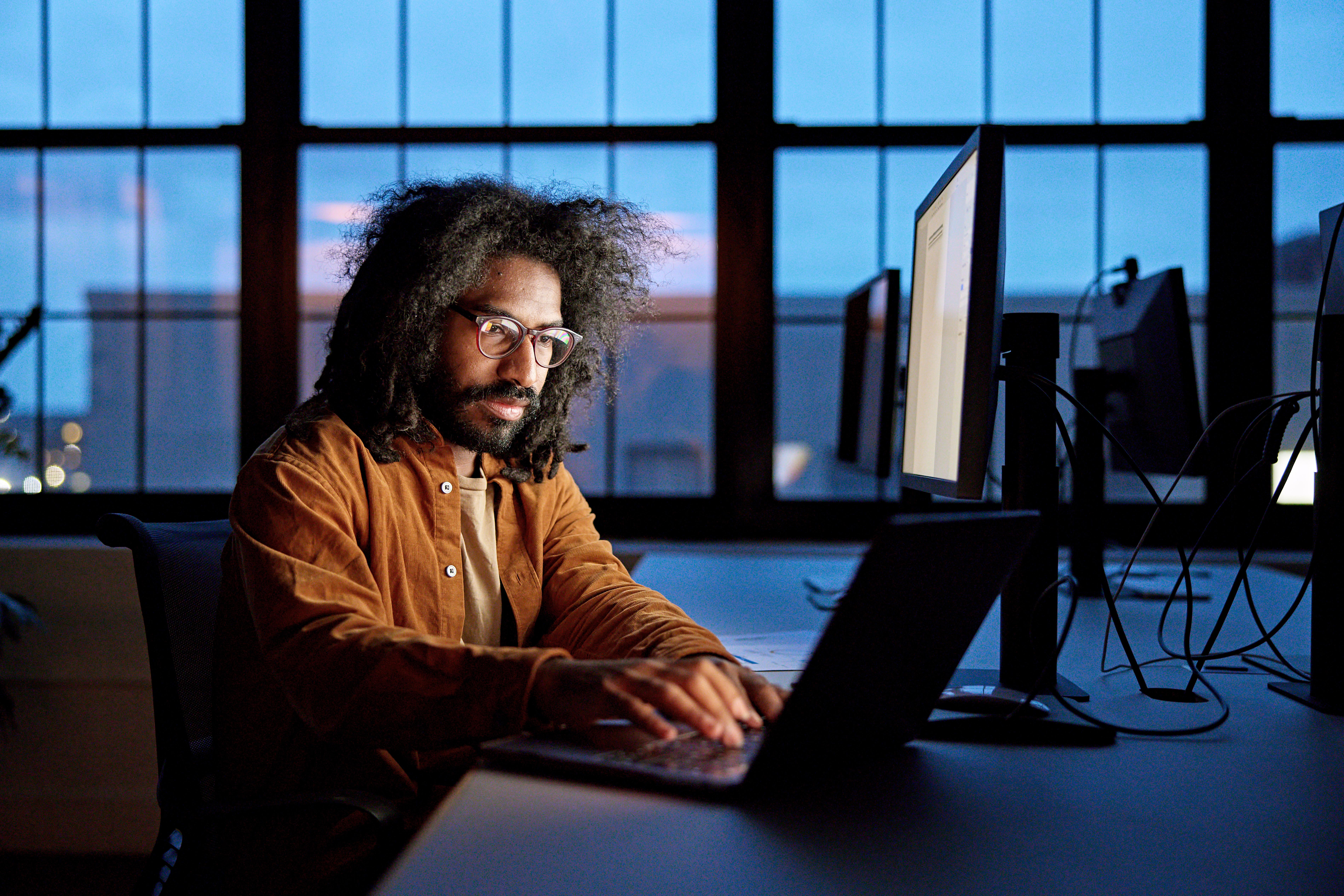 Ein Mann mit Brille und lockigem Haar arbeitet an einem Laptop. Monitore sind hinter ihm. Die Szene hat ein dunkles und blaues Ambiente.