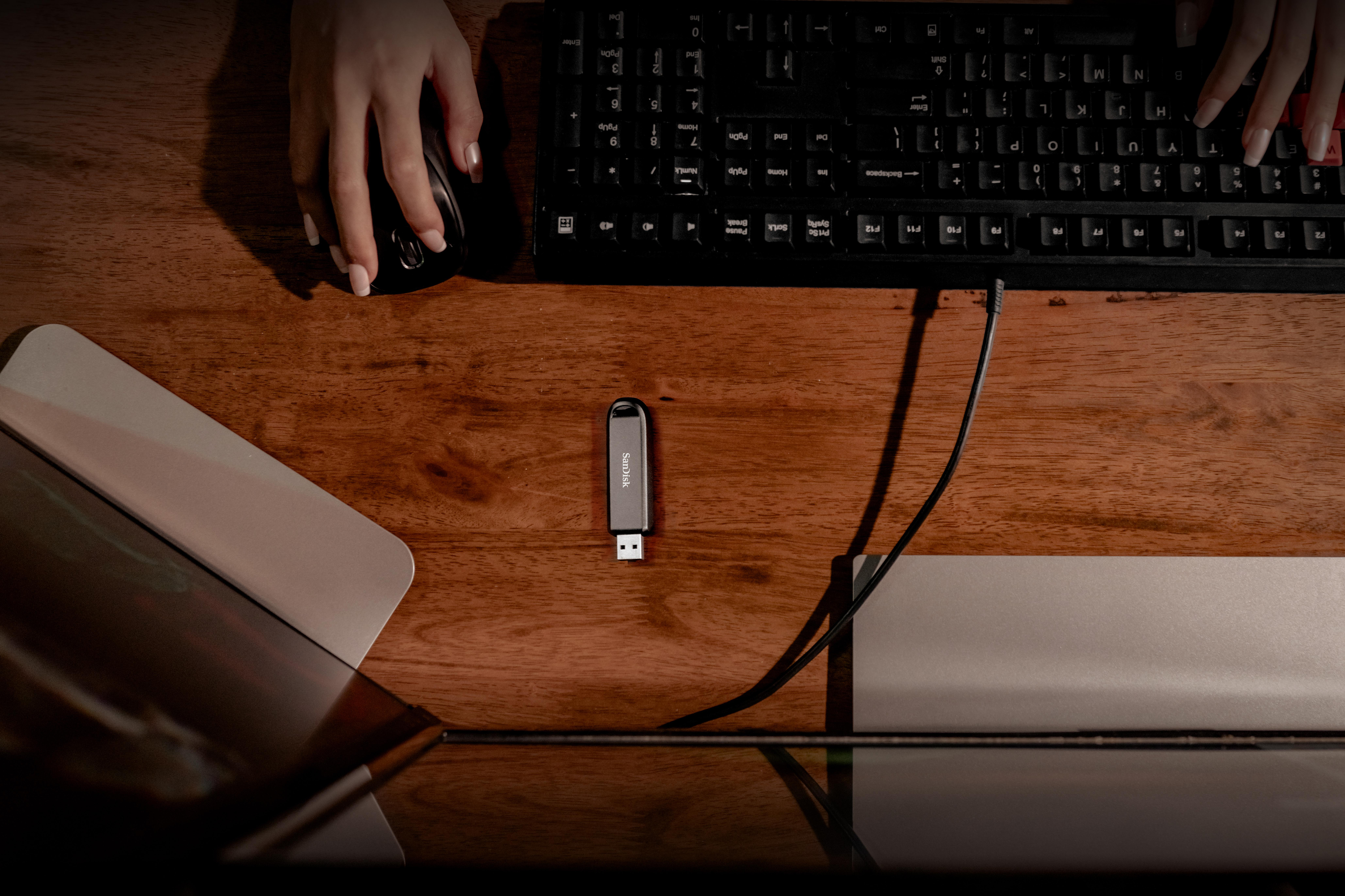 Hände auf einem Schreibtisch mit Tastatur, Maus und USB-Stick. Die Tastatur ist schwarz und der Schreibtisch ist aus Holz.