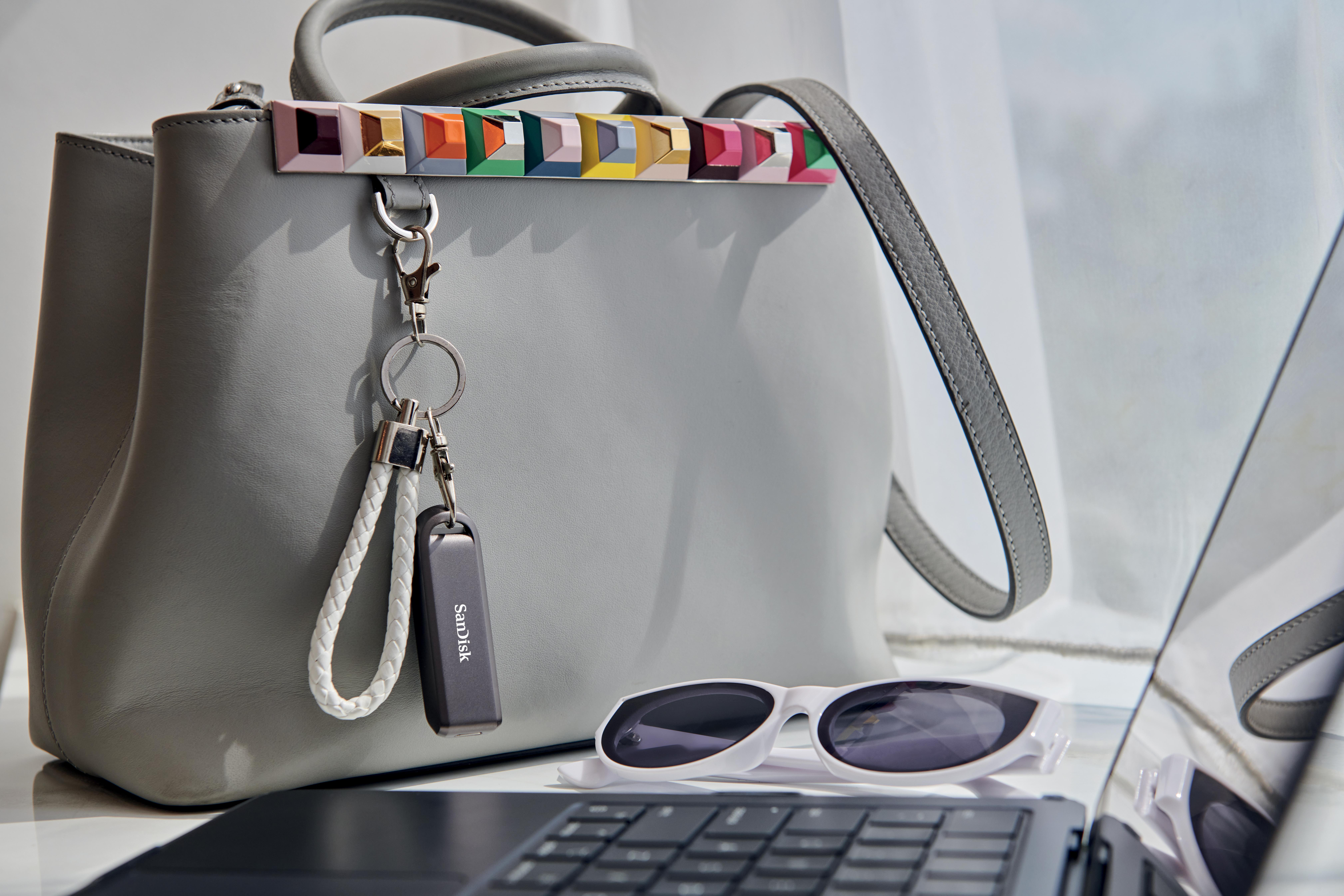 Graue Handtasche mit bunten Akzenten, einem USB-Laufwerk, Sonnenbrille und einem Laptop auf weißer Oberfläche.