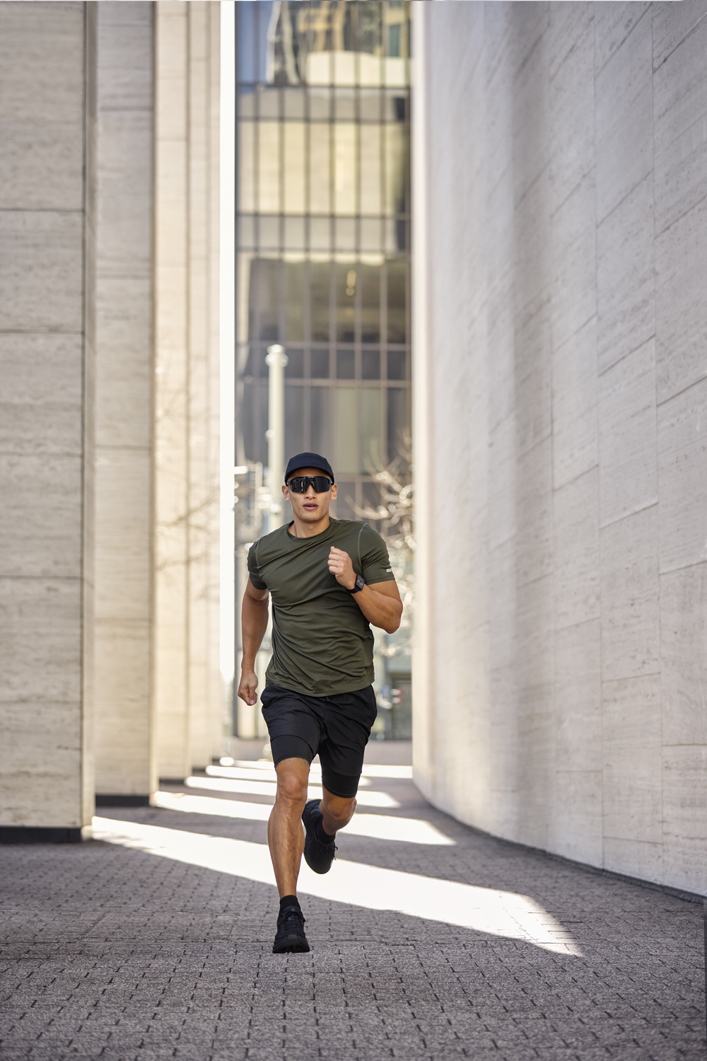 Un hombre corre hacia la cámara. Lleva gorra, gafas de sol, camiseta verde, pantalones cortos negros y zapatos. Edificios al fondo.