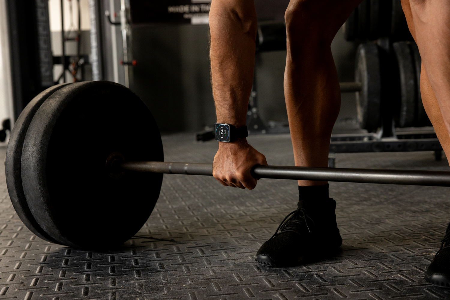 Una persona levanta una barra en un gimnasio. La barra tiene un gran peso. La persona usa un reloj inteligente.