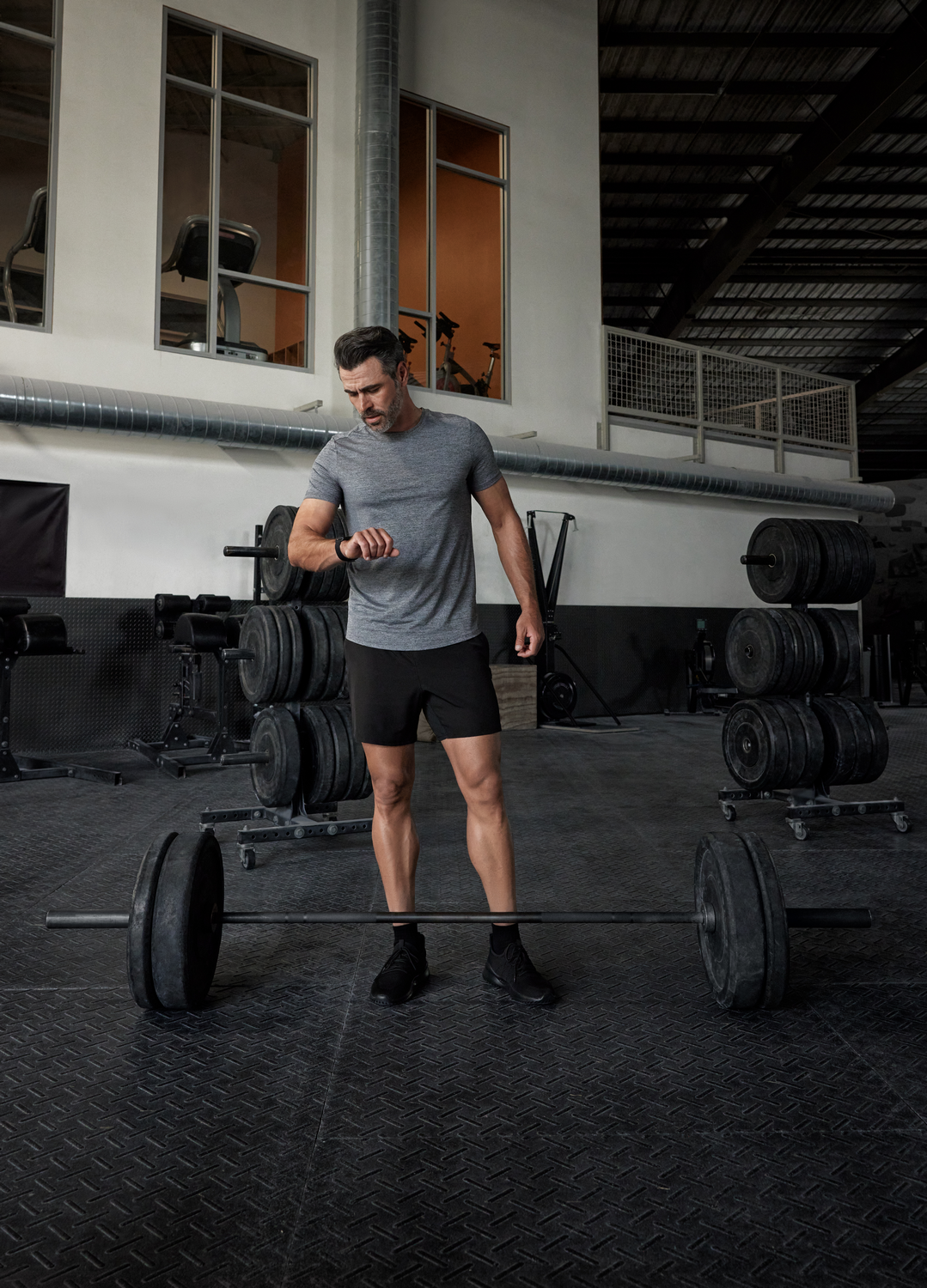 Hombre con ropa de gimnasio mirando reloj, barra delante. Pesas apiladas en estantes. Interior de gimnasio con ventanas.