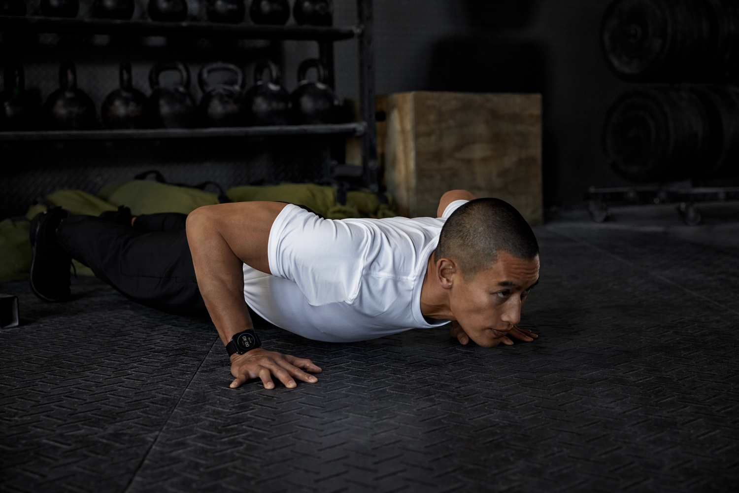 Hombre con camiseta blanca haciendo flexiones en suelo negro. Equipamiento de gimnasio en el fondo. Mira la hora en su reloj.