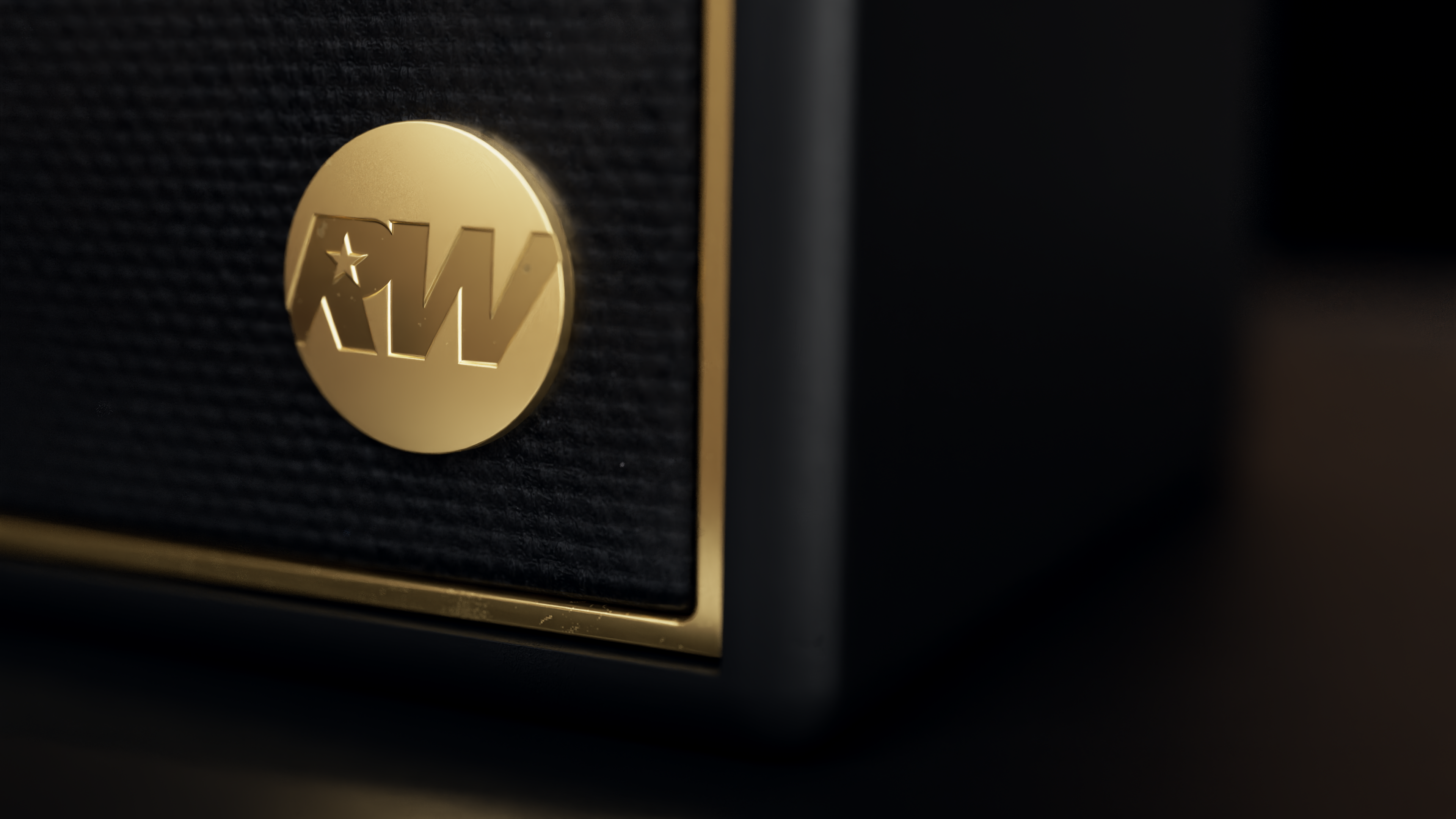 Gros plan sur un haut-parleur noir avec un emblème circulaire doré portant les lettres 'RW' et une étoile. Le fond est noir.