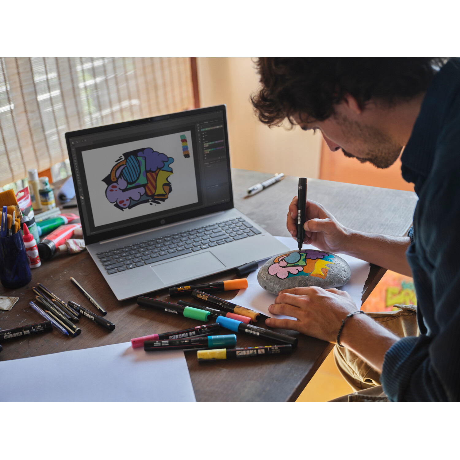 A person is drawing on a rock with markers. A laptop displays a similar design. Markers and art supplies are on the table.