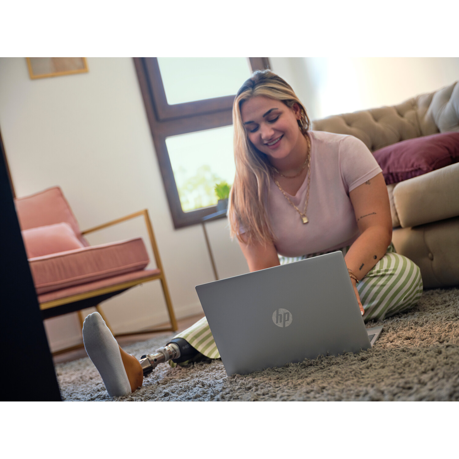 A woman with a prosthetic leg sits on the floor using a laptop. A couch and chair are in the background.