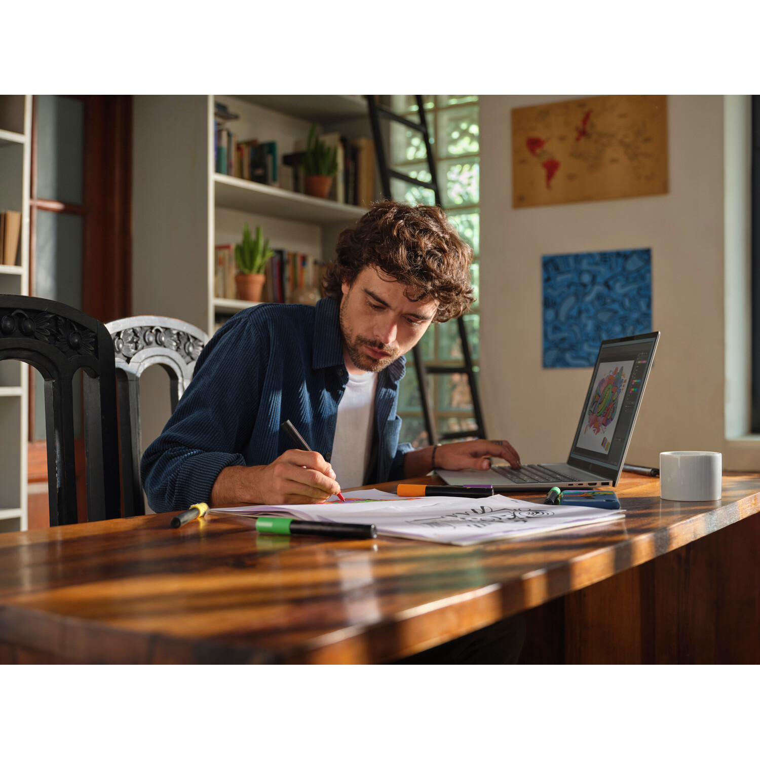 A man sits at a wooden table, drawing. Laptop is open, with a colorful image on the screen. Coffee mug on the table.