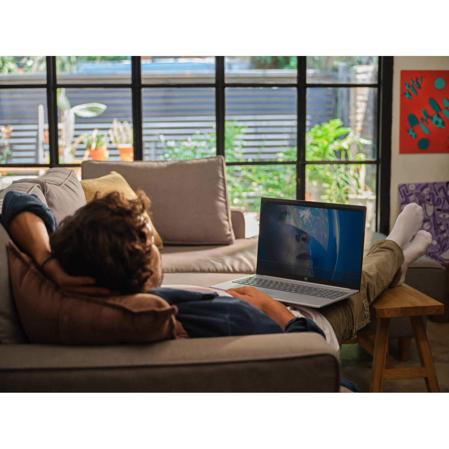 Man relaxing on a couch with laptop. The screen shows an astronaut. The background includes a window and art on the wall.