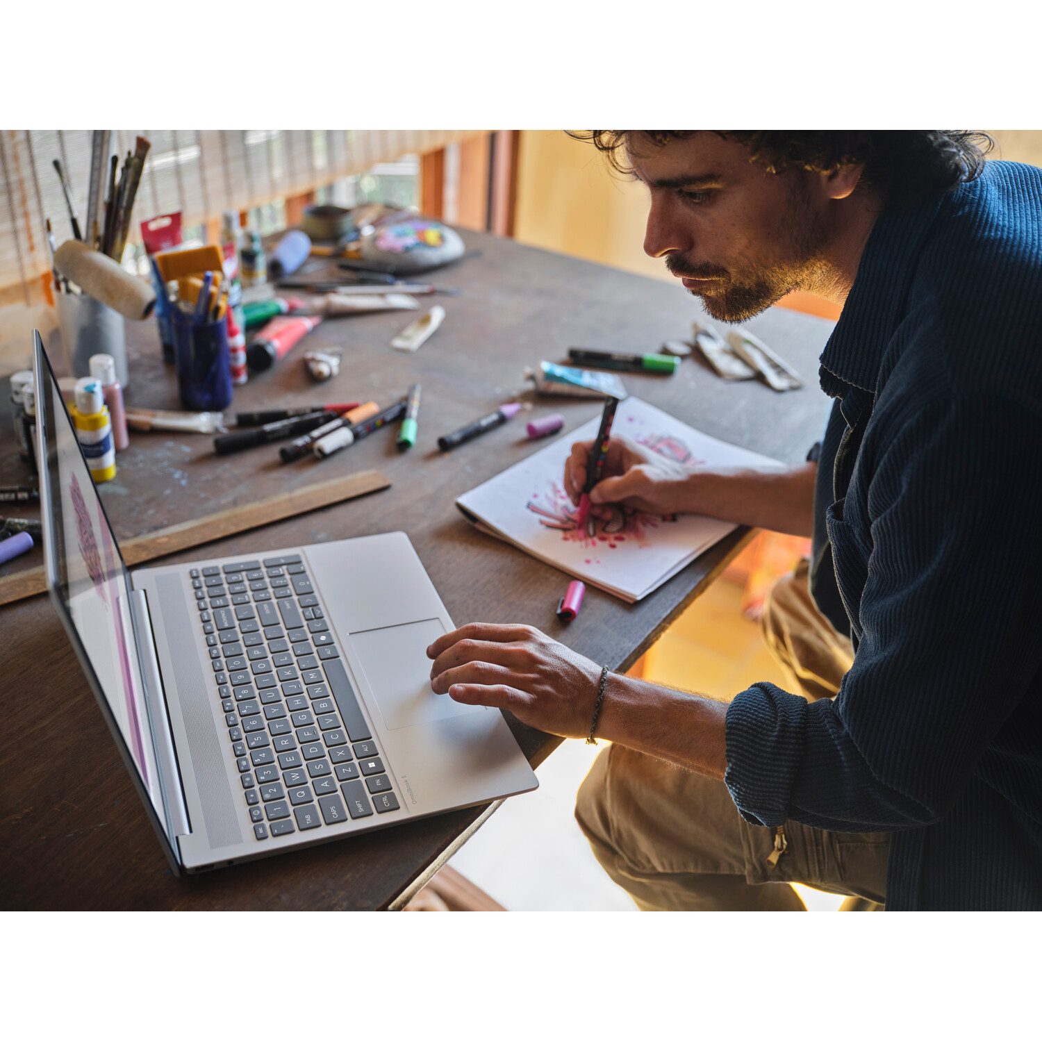 A man draws on paper at a wooden table, laptop open. Art supplies and a paint palette are scattered around. He wears a blue shirt.