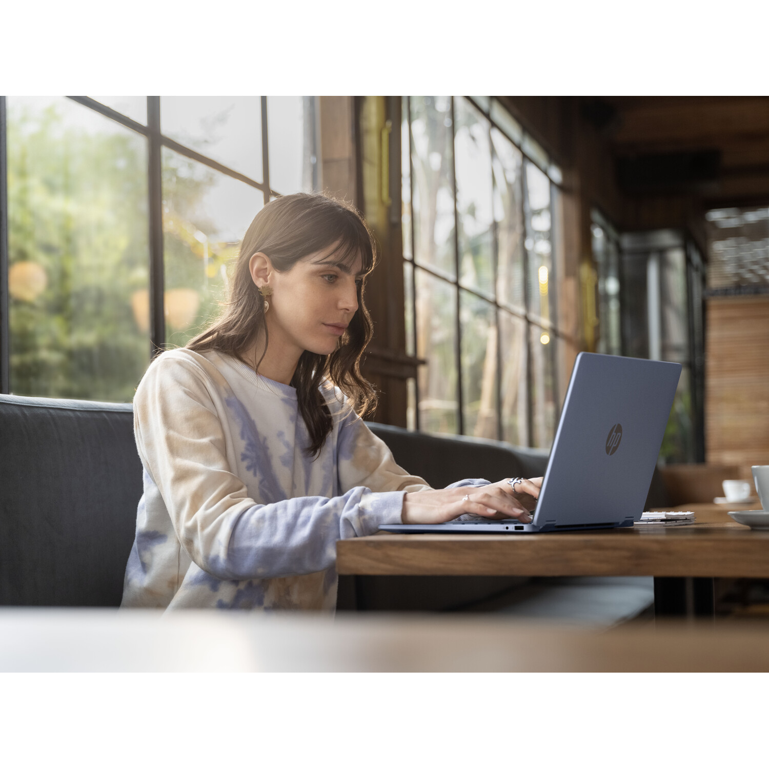 A woman types on a blue HP laptop at a wooden table in a cafe. She wears a tie-dye shirt. Windows are visible in the background.