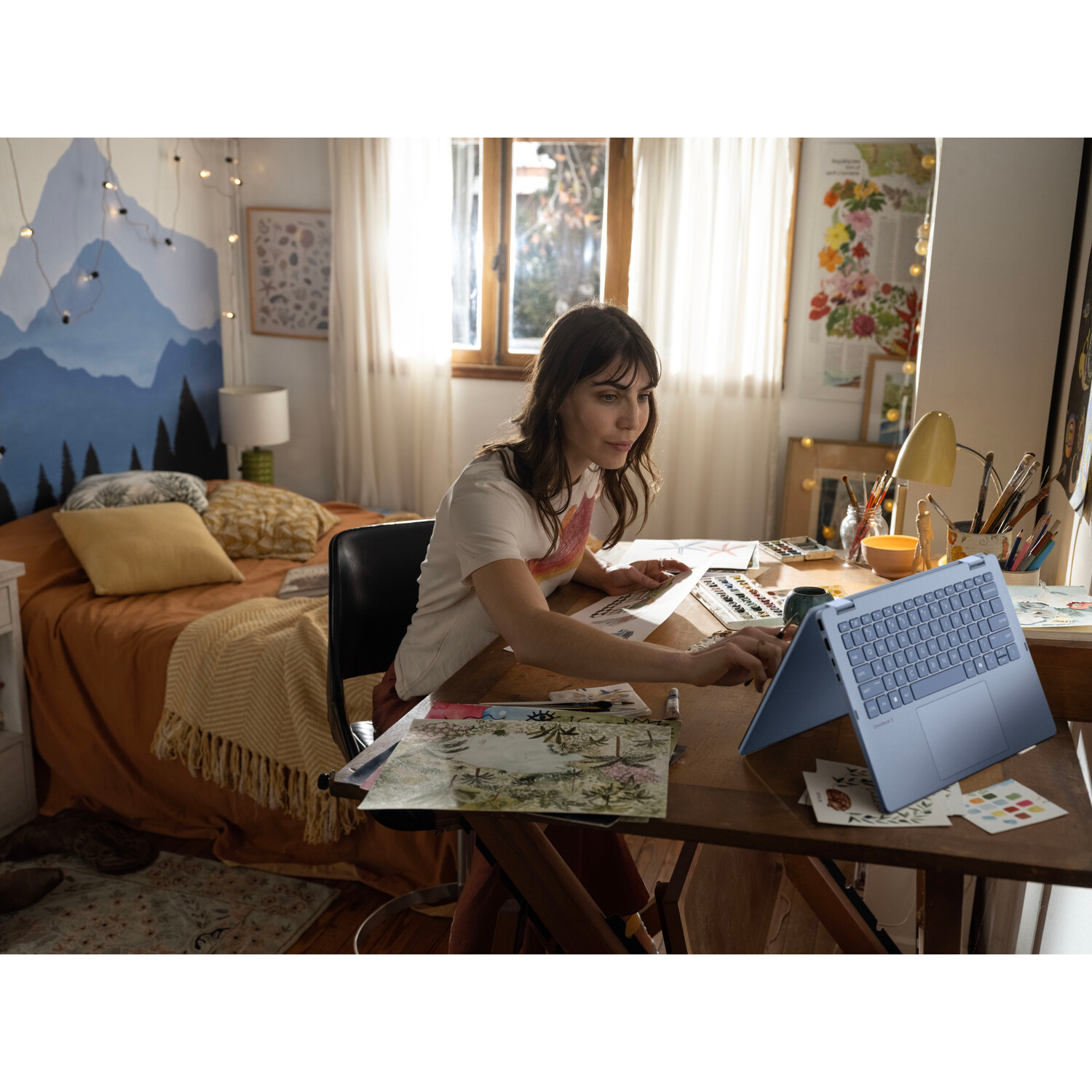 A woman sits at a desk with a laptop. She works on art projects in a bedroom. A bed is in the background. The room has art on the walls.