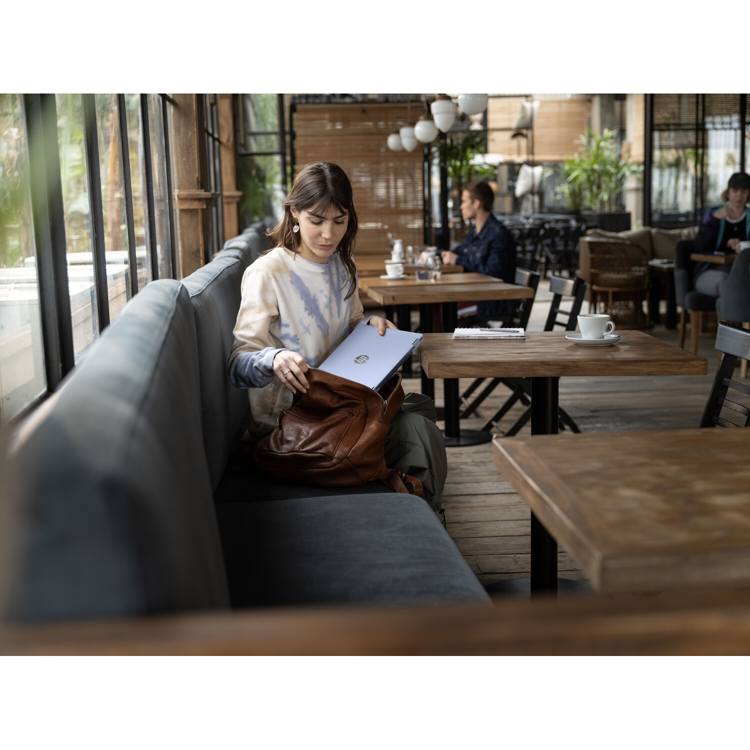 Woman in cafe, placing laptop in brown bag. Other people sit at tables. Cafe has large windows and wooden tables.