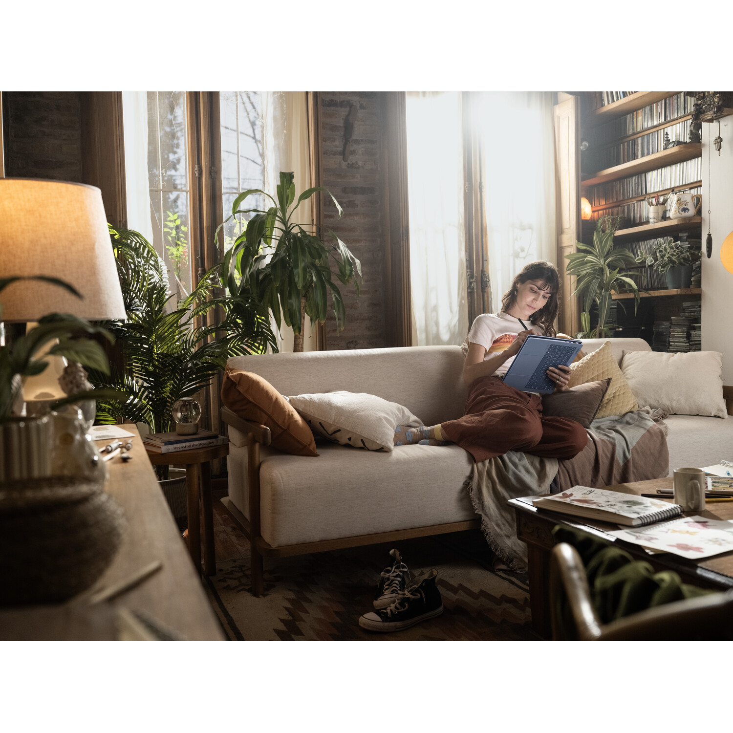 A woman sits on a couch, using a blue tablet. She's in a cozy living room with plants, a bookshelf, and natural light.