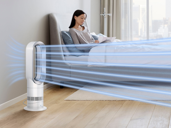 Una mujer lee en la cama. Un ventilador blanco sopla aire. Una corriente azul fluye sobre la cama. La habitación tiene un esquema de color claro.