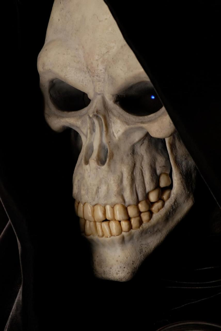 Nahaufnahme eines Totenkopfgesichts mit schwarzer Kapuze. Es hat Knochenfarbe, dunkle Augenhöhlen und sichtbare Zähne. Ein blaues Licht in einem Auge.